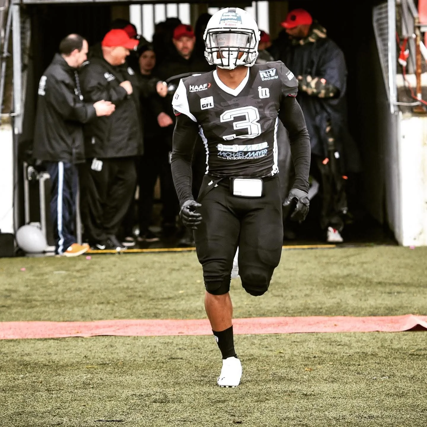 American football player wearing jersey number 3, running onto the field through a tunnel with team members and staff in the background.