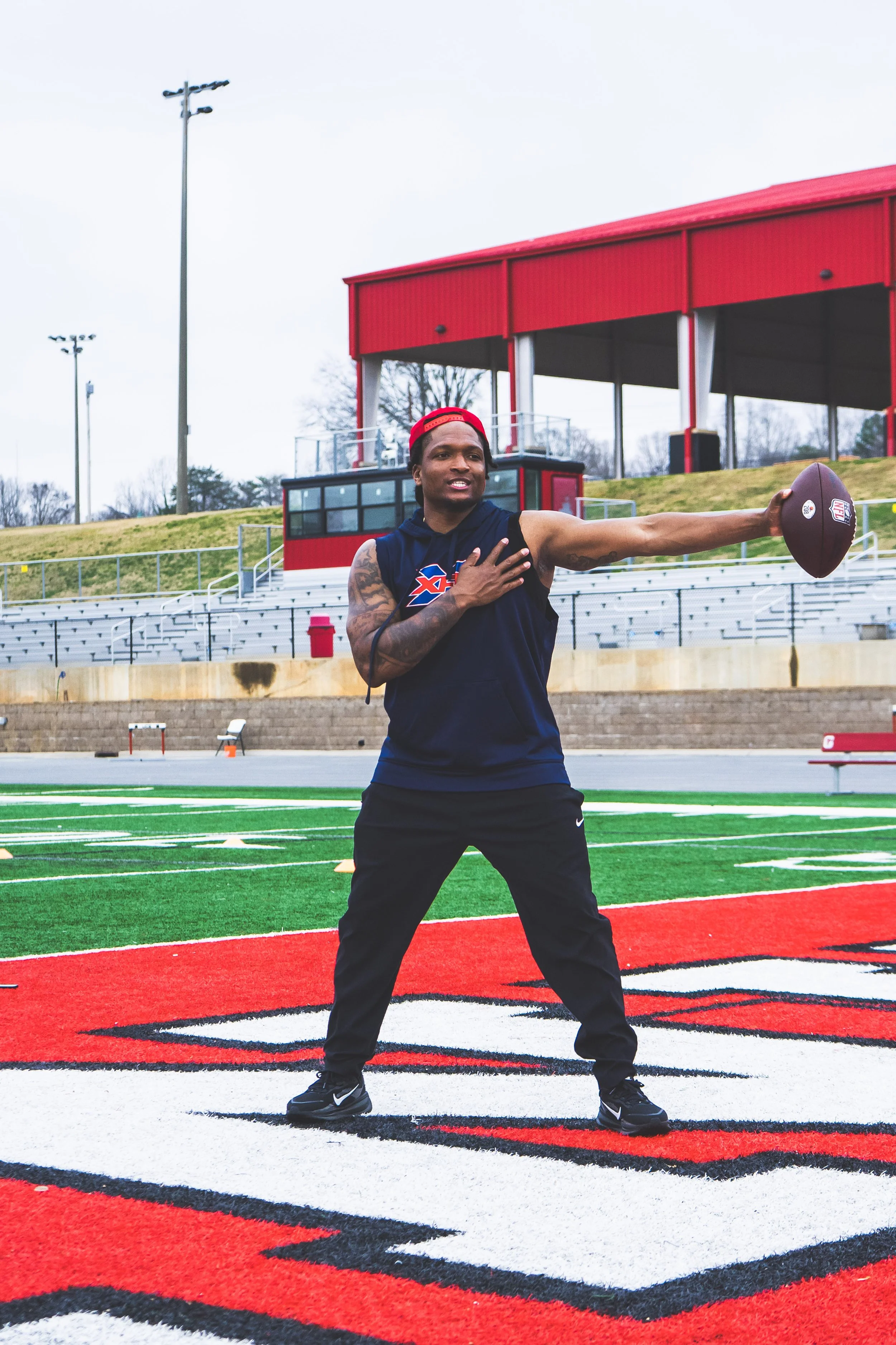 A man on a football field holding a football, wearing athletic clothes and a red cap backward, with his right hand on his chest and his left arm extended holding the football, on a sports field with red, green, and black markings and stadium seating 