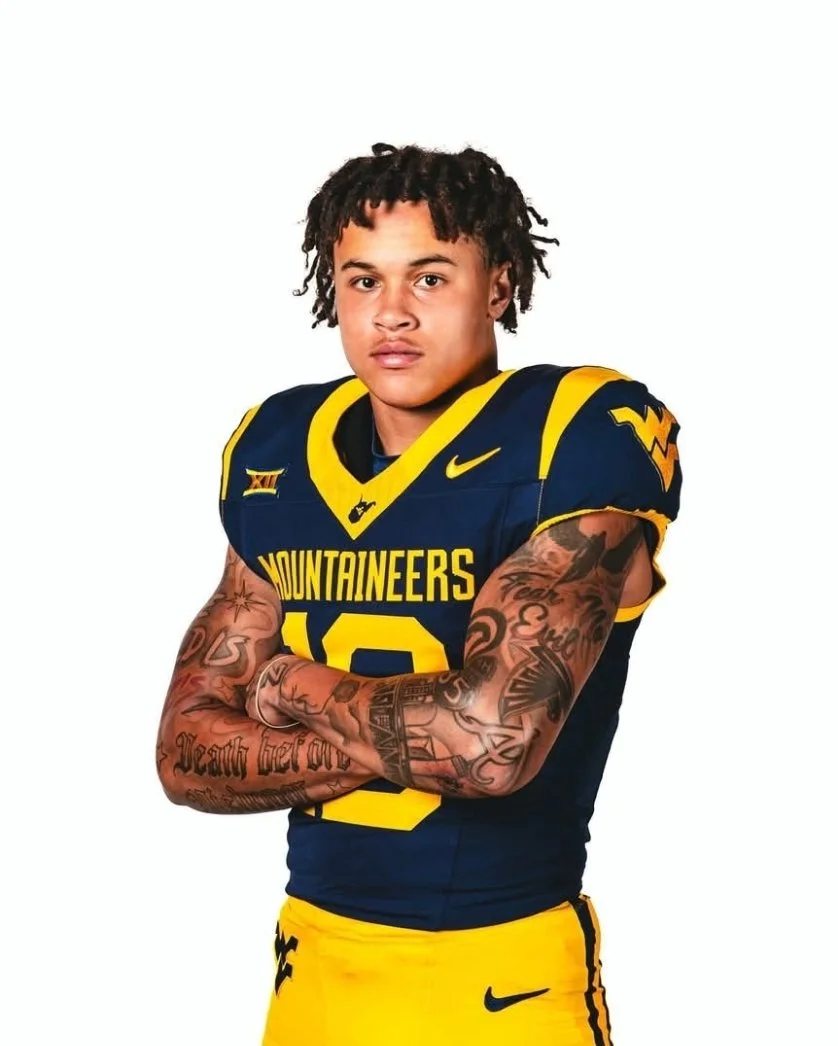 A young man in a West Virginia Mountaineers football uniform with arms crossed, tattoos on his arms, and a neutral expression