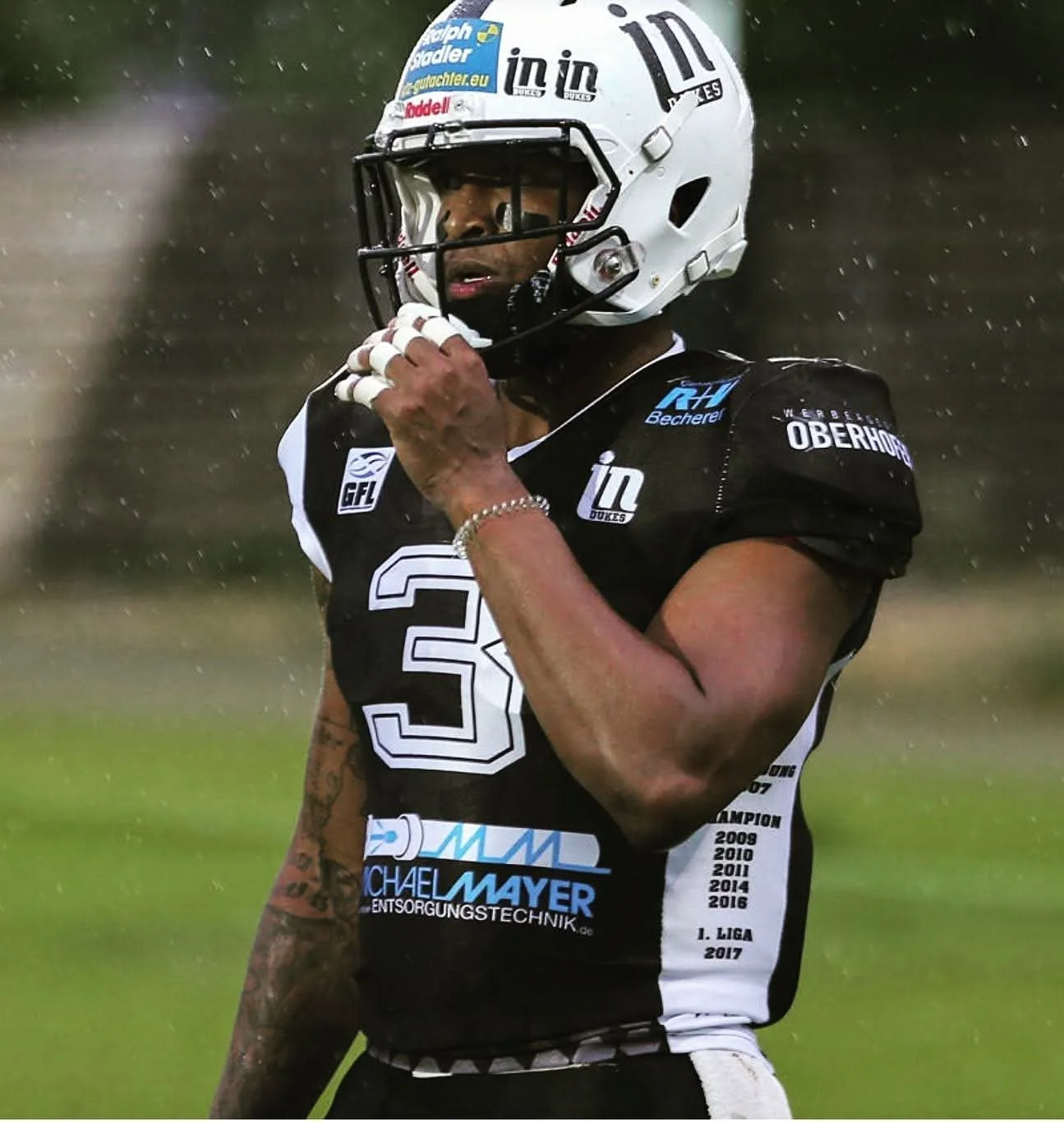 An American football player wearing a white helmet and black jersey with the number 3, standing on a football field in the rain, adjusting his helmet strap.