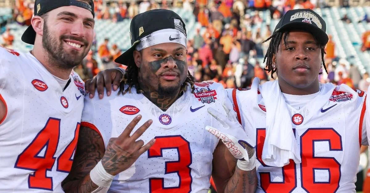 Three football players in white uniforms with orange and blue accents, standing together on the field. They are wearing caps, and the background shows a stadium filled with spectators.