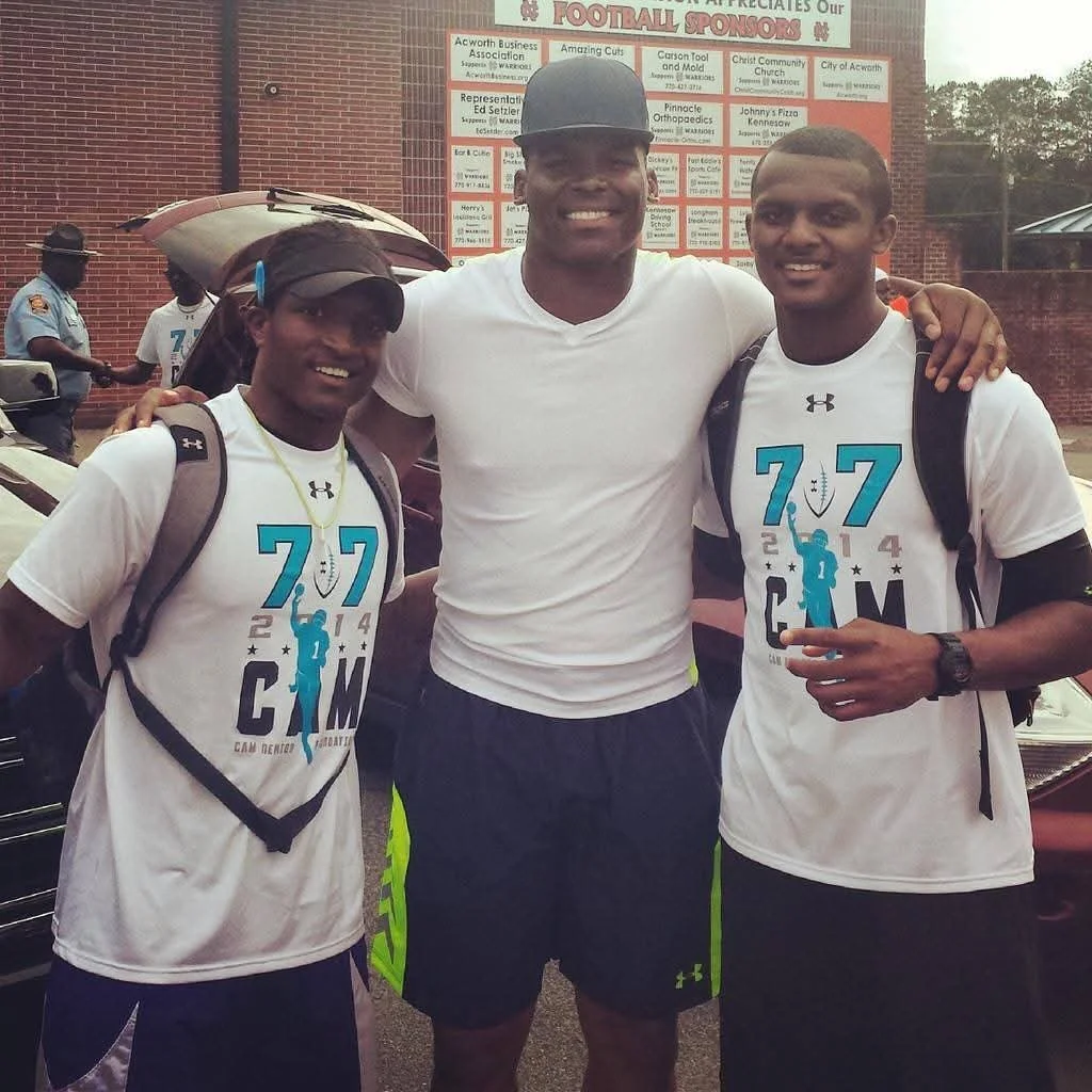 Three young men smiling with arms around each other, two wearing white T-shirts featuring the 2014 CAM football event logo, and one in the middle wearing a gray cap and white shirt. They are outdoors near a brick building with a large sign and some p