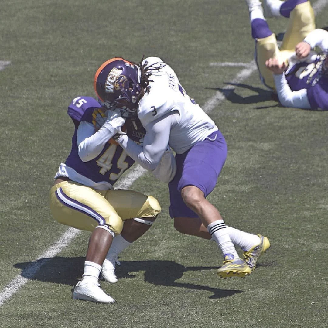 Two football players in white and purple uniforms are engaged in a tackle on a football field, with other players in the background.