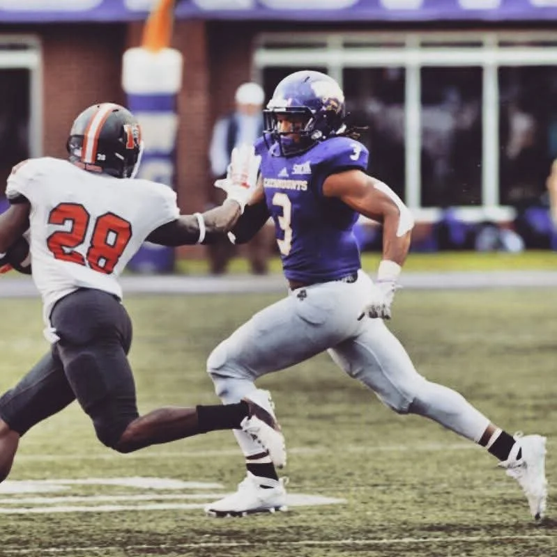 Two football players in action on the field; the player in the white and black uniform with the number 28 is attempting to block or tackle the player in the purple and white uniform with the number 3.