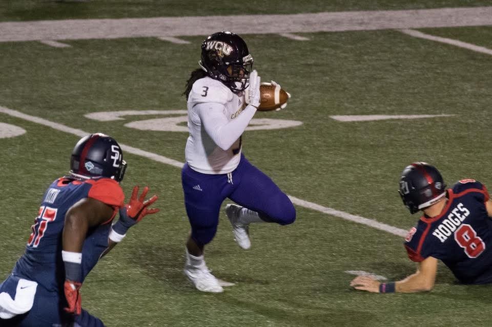 A football player holding the ball running past two defenders on the field.