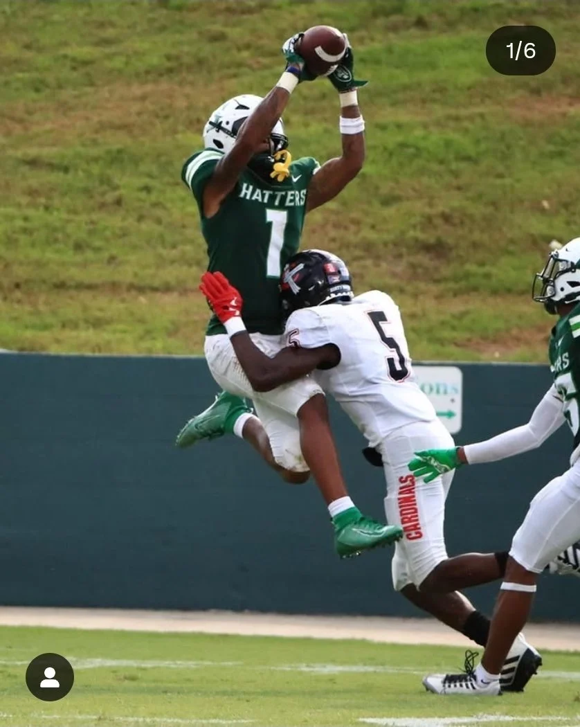 American football players in action; one player in a green jersey with 'HATTERS' and the number 1 is jumping to catch the ball, another player in a white jersey with the number 5 is attempting to tackle him, on the field with a grassy background.