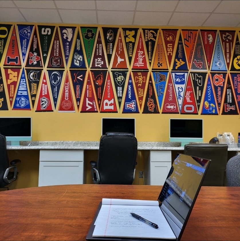 Wall decorated with college sports team banners, and a workspace with a laptop, notebook, and pen on a wooden table.
