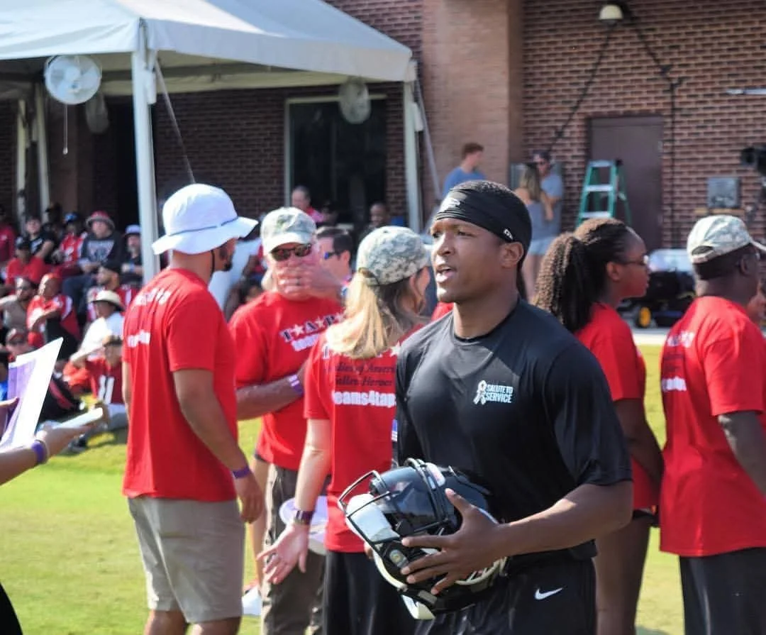 Young man in sportswear holding a football helmet on a football field, surrounded by people in red shirts during a community event or gathering.