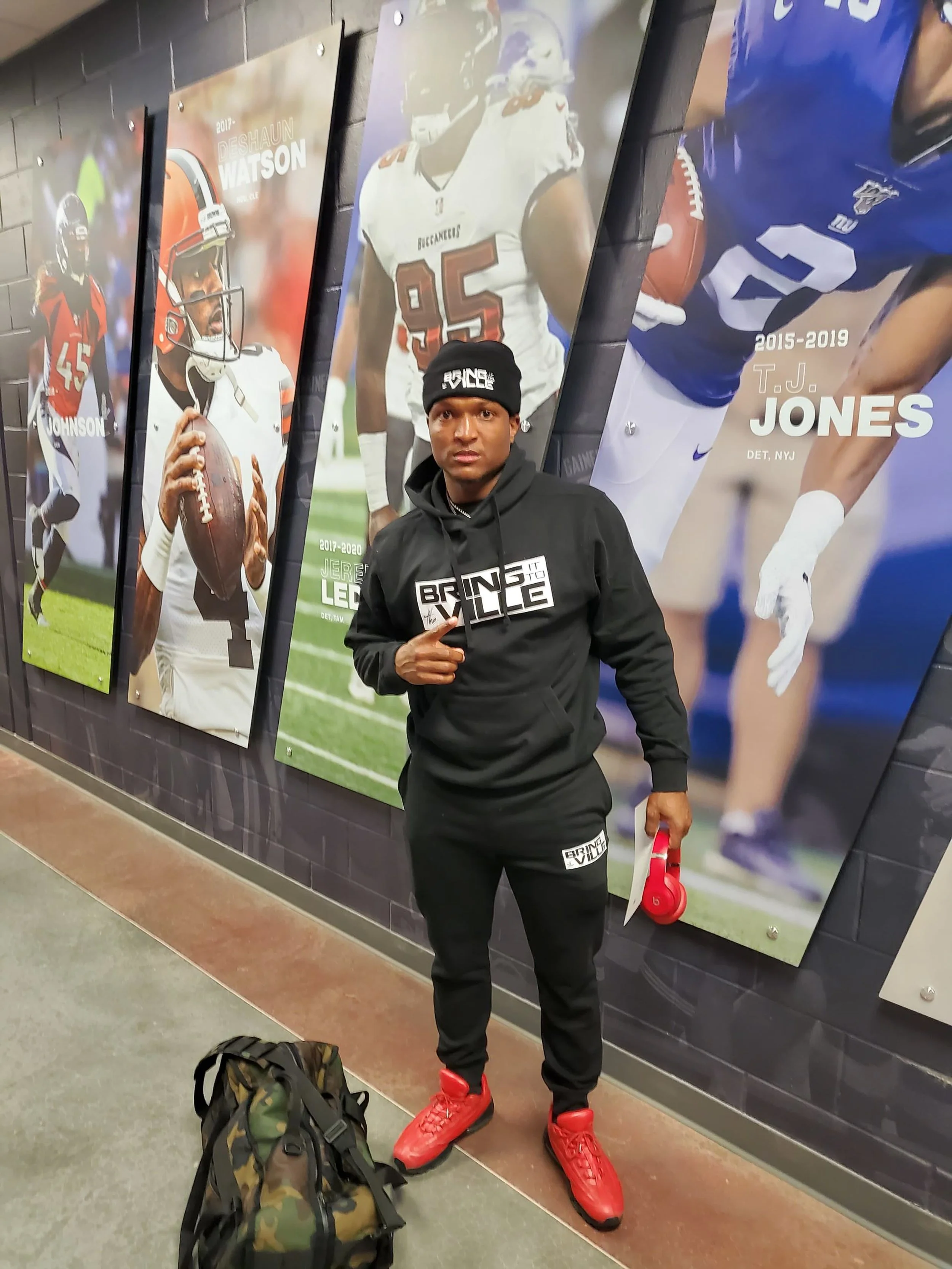 A man wearing a black hoodie, black pants, and red sneakers standing in front of large football posters on a wall.