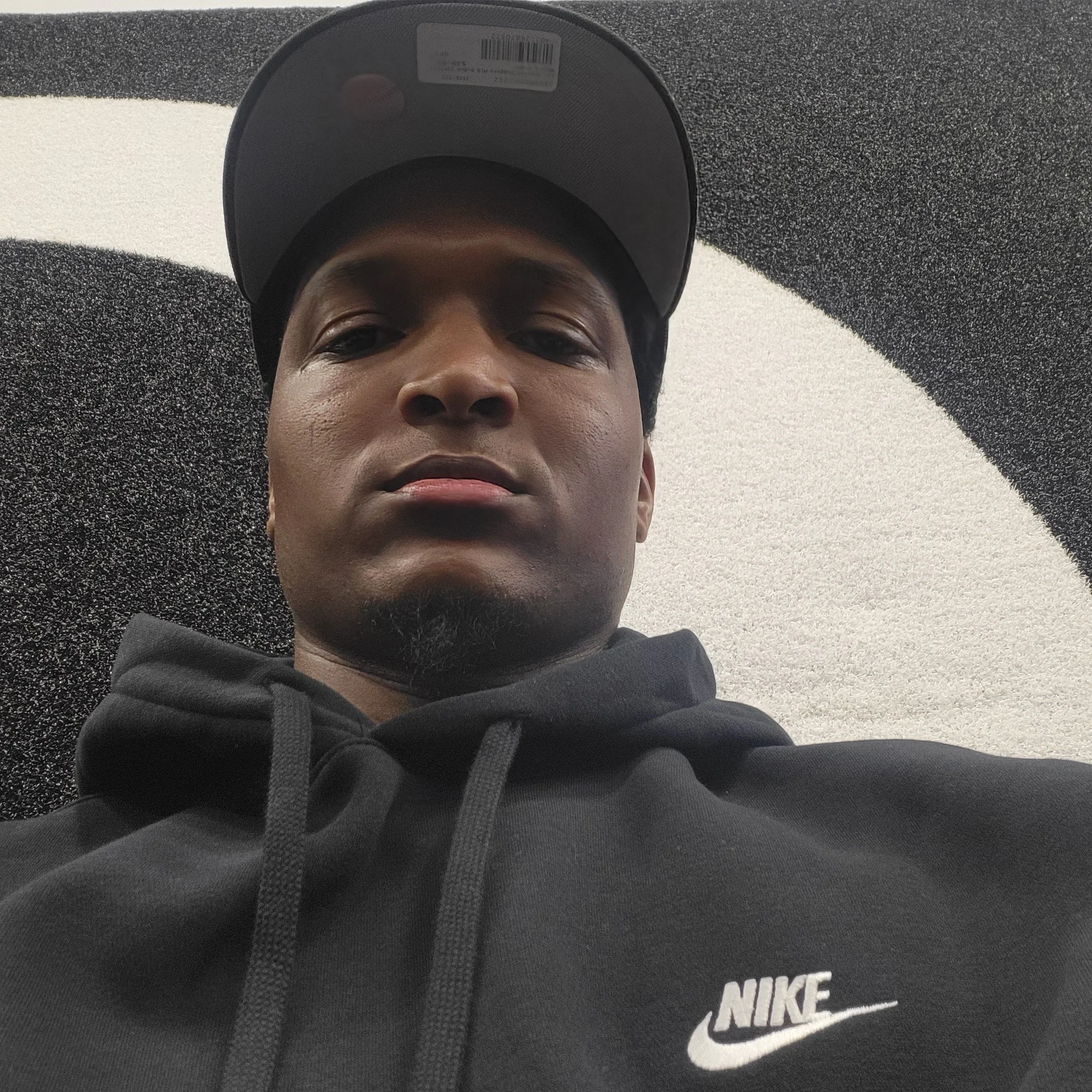 A young man wearing a black Nike hoodie and a black baseball cap, taking a selfie from a low angle indoor against a black and white wall.