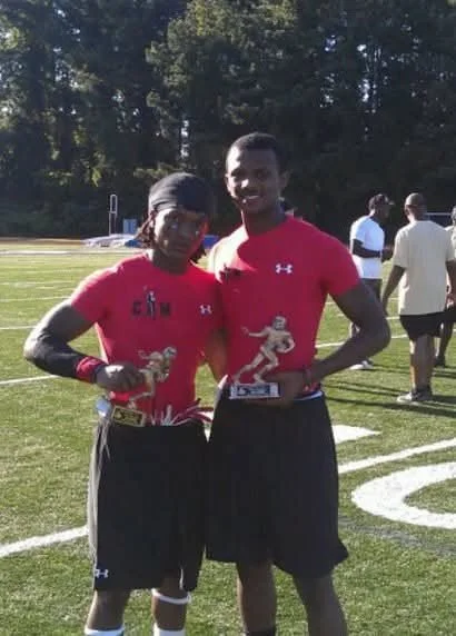 Two men in red athletic shirts holding trophies, standing on a football field with other people in the background