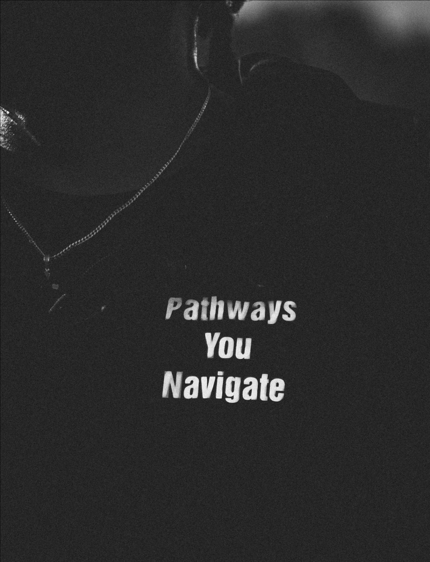 Close-up of a person wearing a necklace and a dark t-shirt with the partially visible text 'Pathways You Navigate,' in a dark setting.