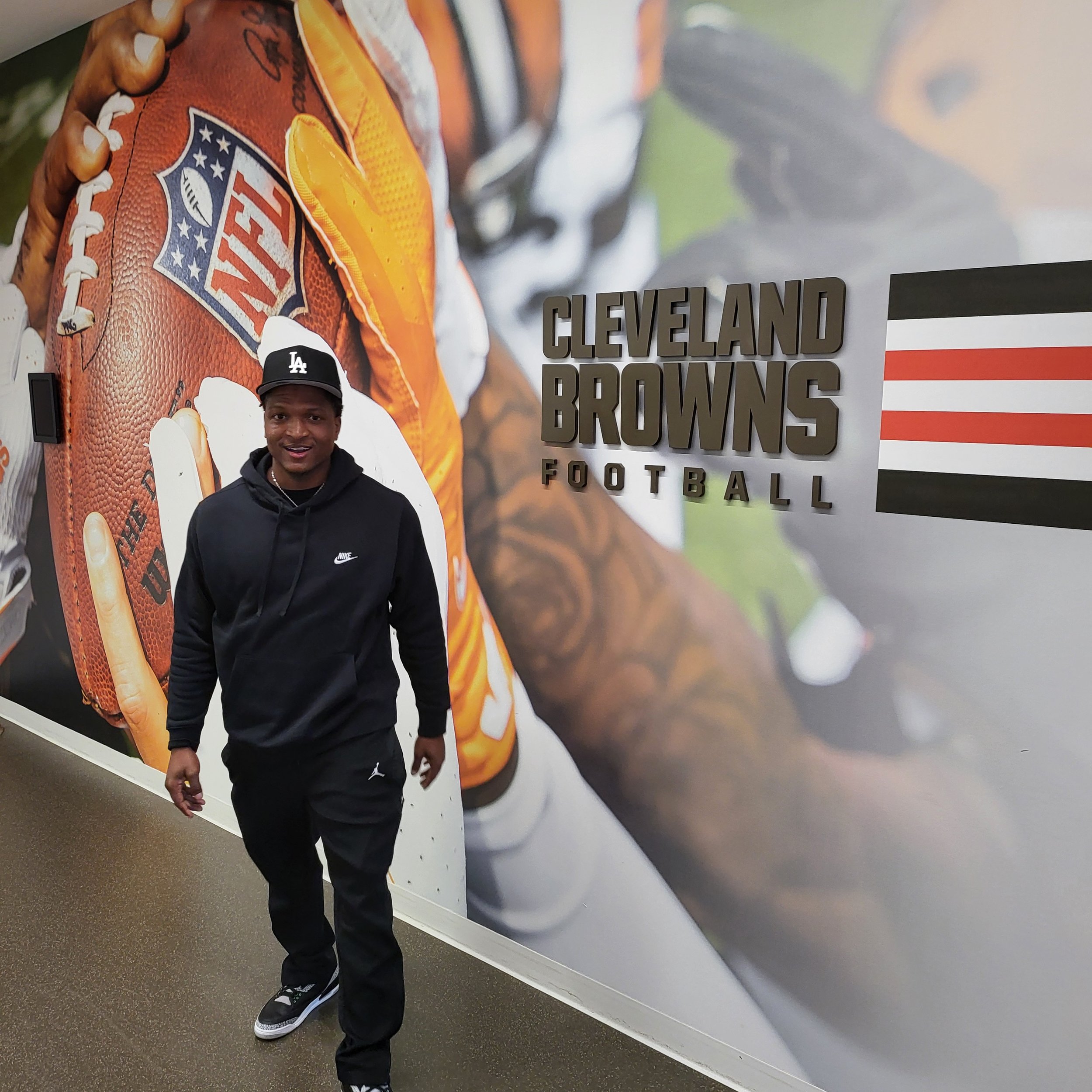 A young man wearing a black hoodie with a Nike logo, black pants, and sneakers stands in front of a large Cleveland Browns football poster. The poster features a football, the NFL shield logo, and the Cleveland Browns team name.
