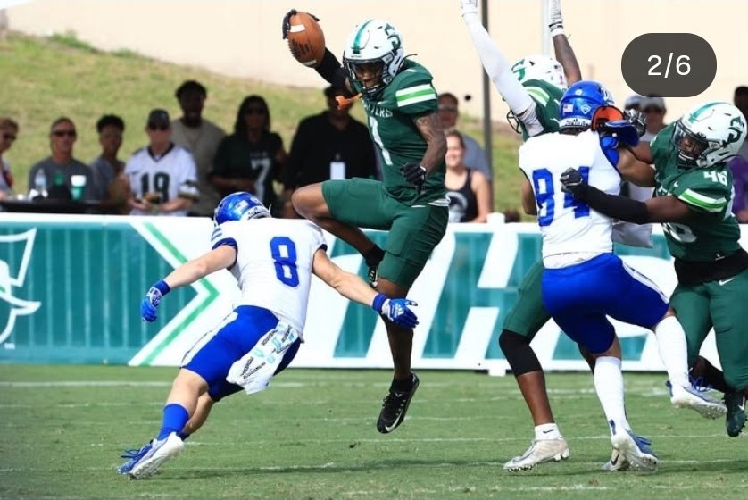 A football game in progress with players in green and white uniforms. One player in green is jumping in the air with the ball, while opponents in white and blue attempt to stop him. Spectators are watching from the background.