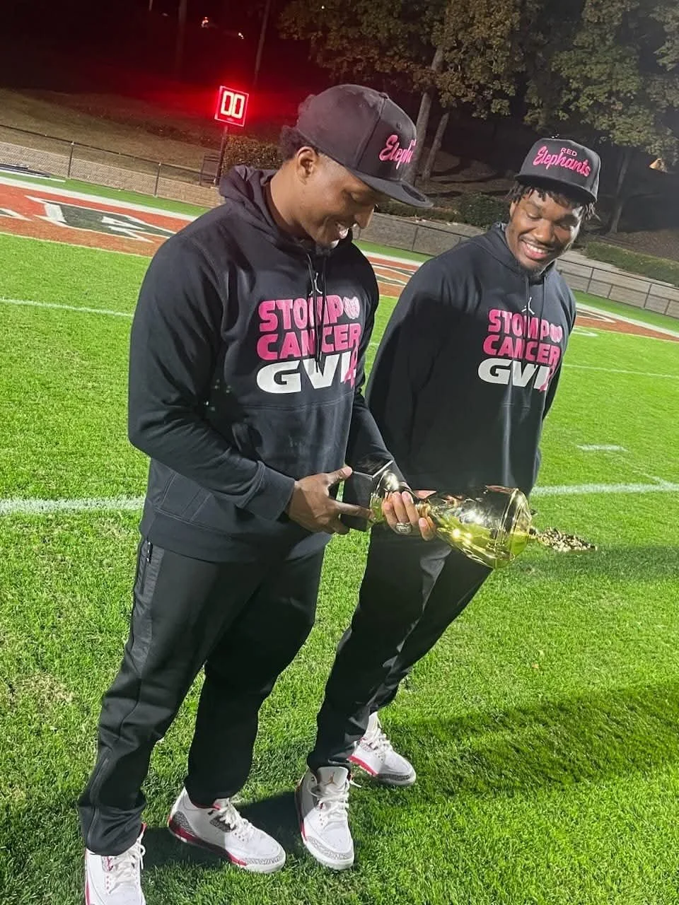 Two young men wearing black hoodies and caps with pink text, standing on a football field at night, pouring a beverage from a bottle into a large glass vase, smiling and enjoying the moment.