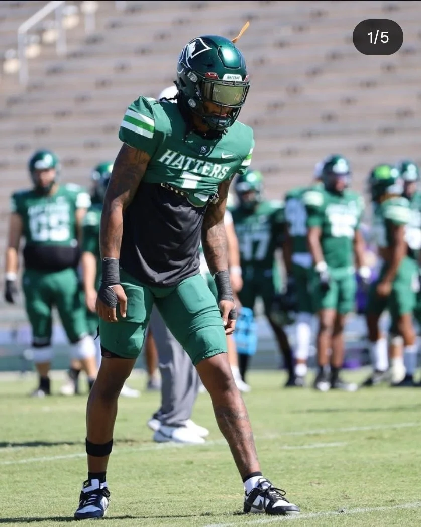 Football player in a green uniform and helmet, standing on the field during a game, with teammates in the background.