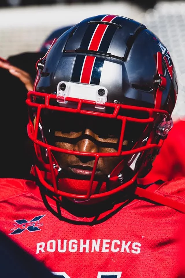 A football player wearing a red jersey with the word 'ROUGHNECKS' and a logo on it, a dark blue helmet with red and white stripes and a faceguard, and sunglasses, during a game or practice.