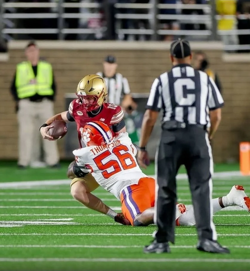 A football player in a red and gold uniform running with the ball while a player in a white and orange uniform tackles him on the field during a game, with an official observing nearby.
