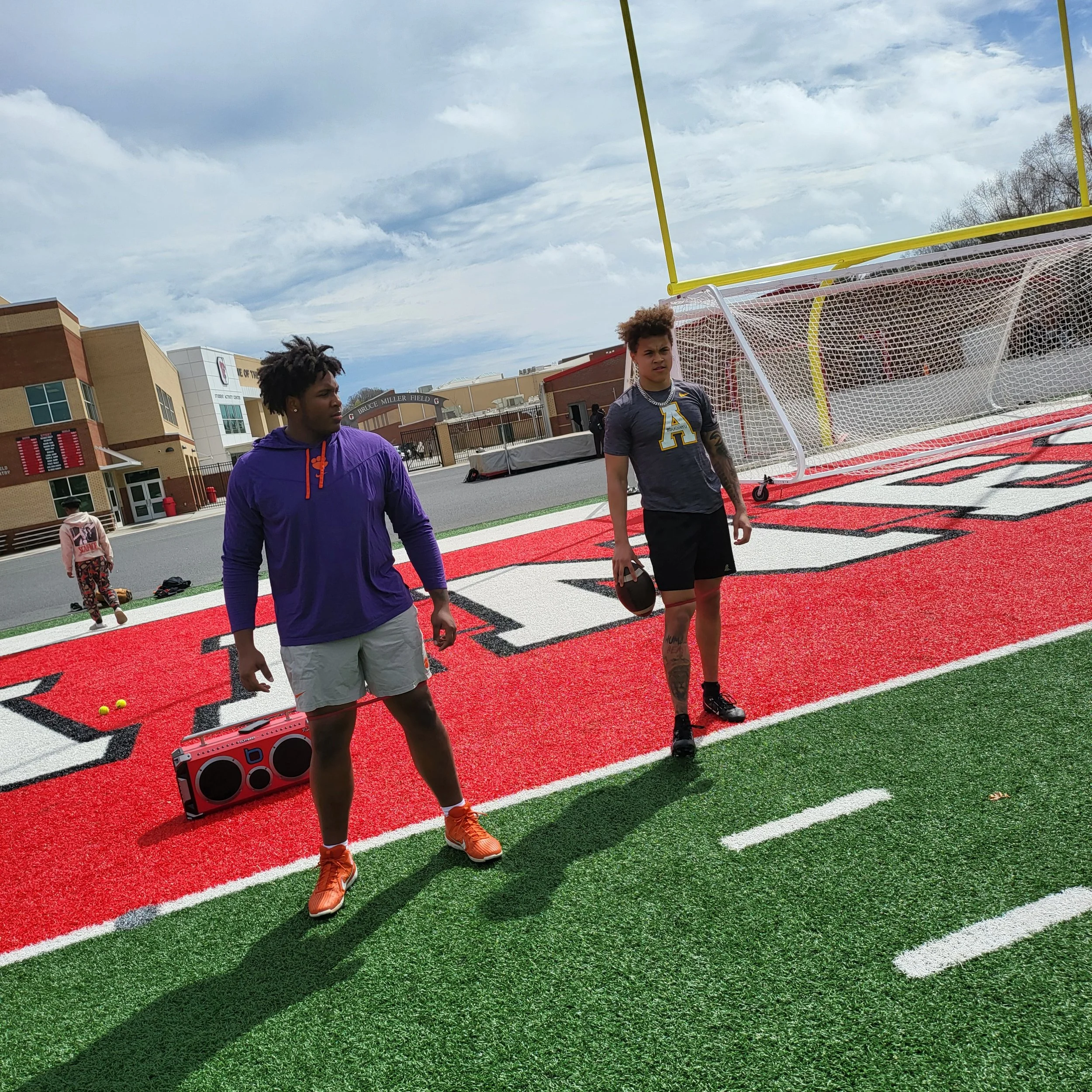 Two young men standing on a football field near the goalpost, one holding a football, on a sunny day with some clouds, with school buildings in the background.