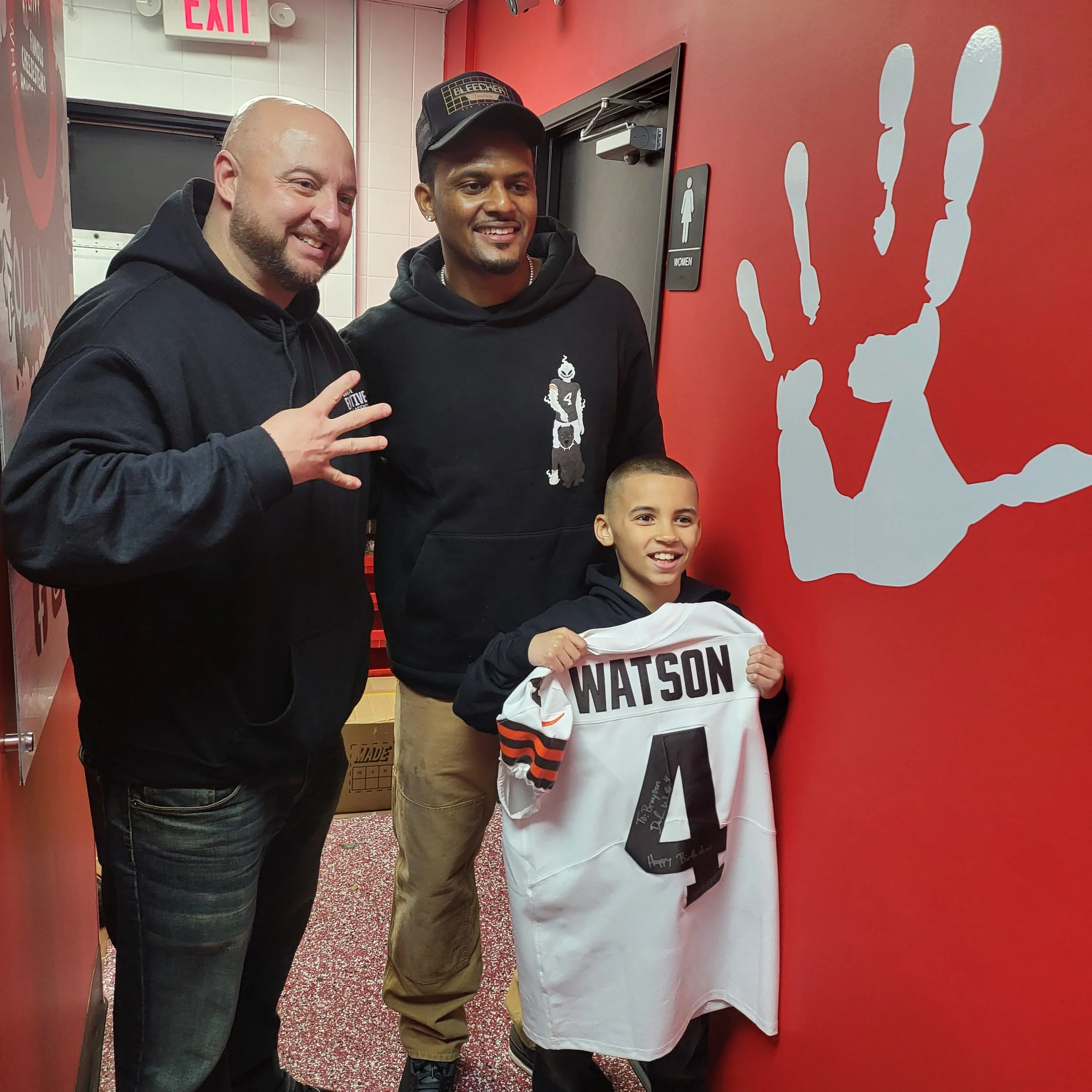 Two men and two boys standing in front of a red wall with white silhouettes of football players. The man on the left is smiling, wearing a black hoodie, and making a rock sign with his hand. The man in the middle is smiling, wearing a black hoodie an