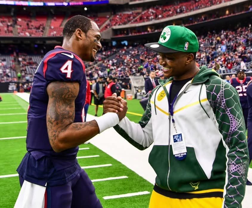 Two athletes are smiling and shaking hands on a football field before a game. One is wearing a navy blue uniform with the number 4, and the other is wearing a green cap and a white jacket with green and purple accents.
