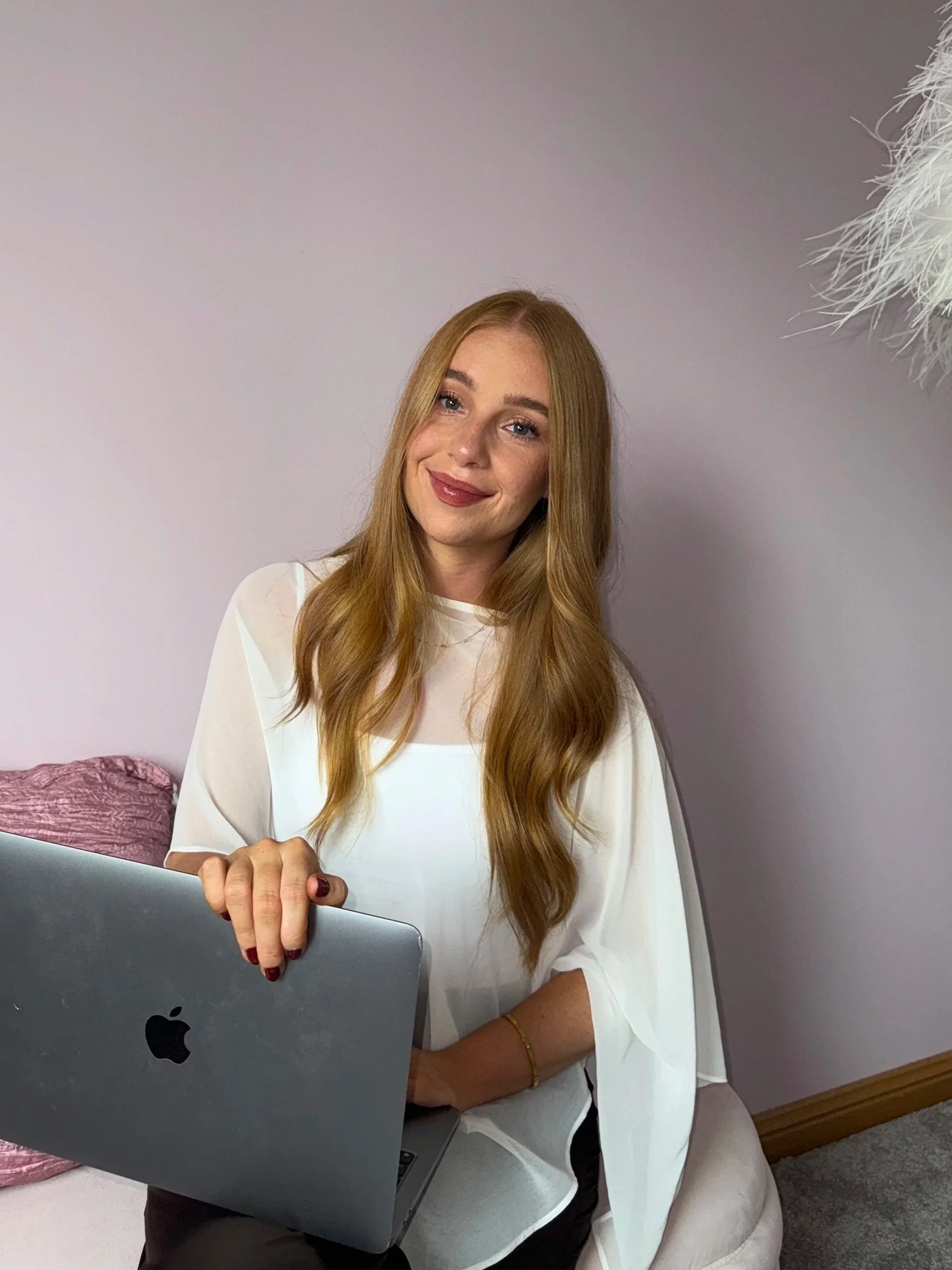 A woman with long, wavy red hair and fair skin sitting on a light-colored chair, holding a silver MacBook laptop, smiling slightly, wearing a white blouse, gold bracelet, in a room with a pink pillow and a large white feathered decoration on the wall.