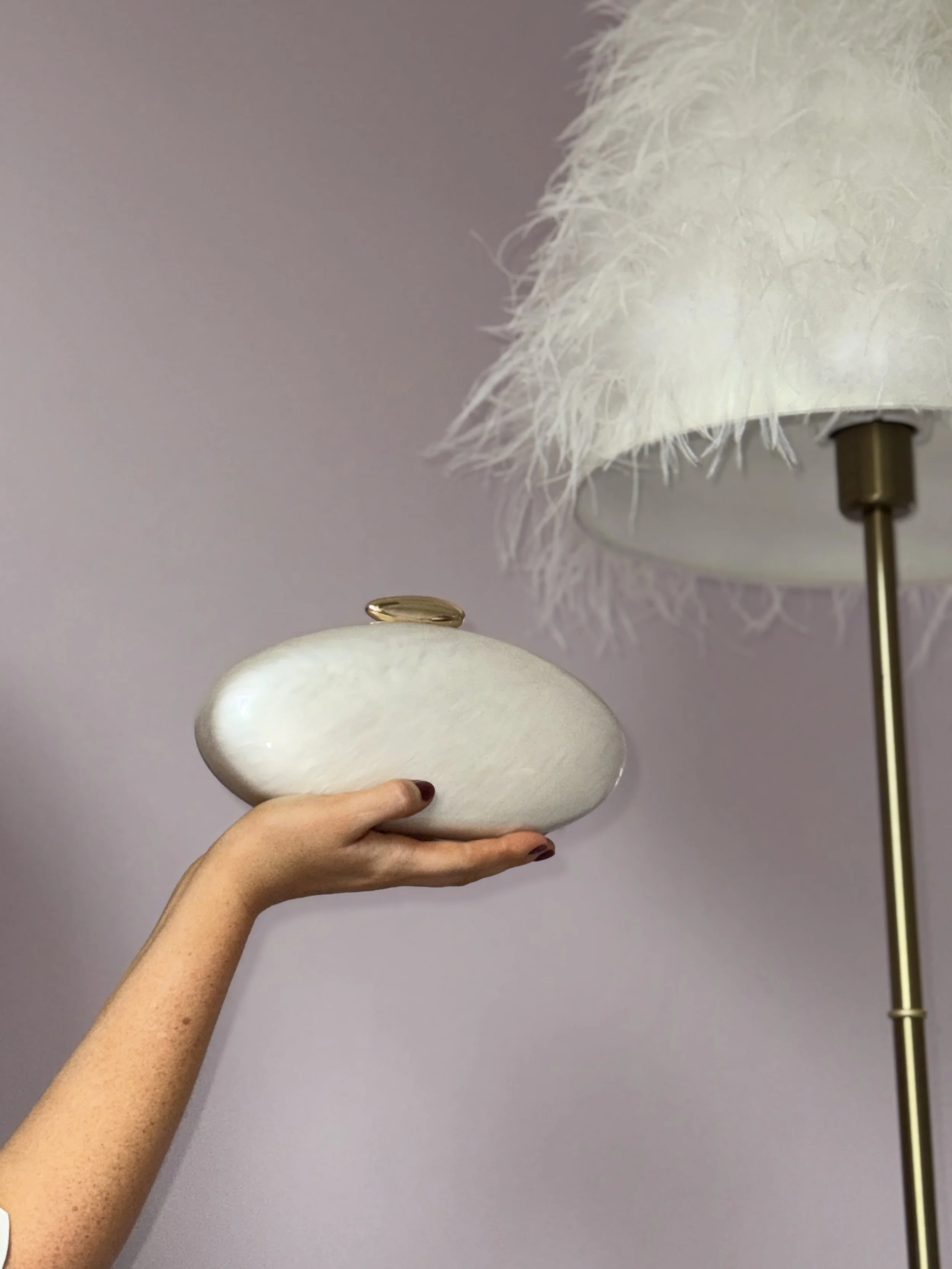 A person's hand holding a white, fluffy, oval-shaped lamp base near a white feather lamp shade on a brass stand.