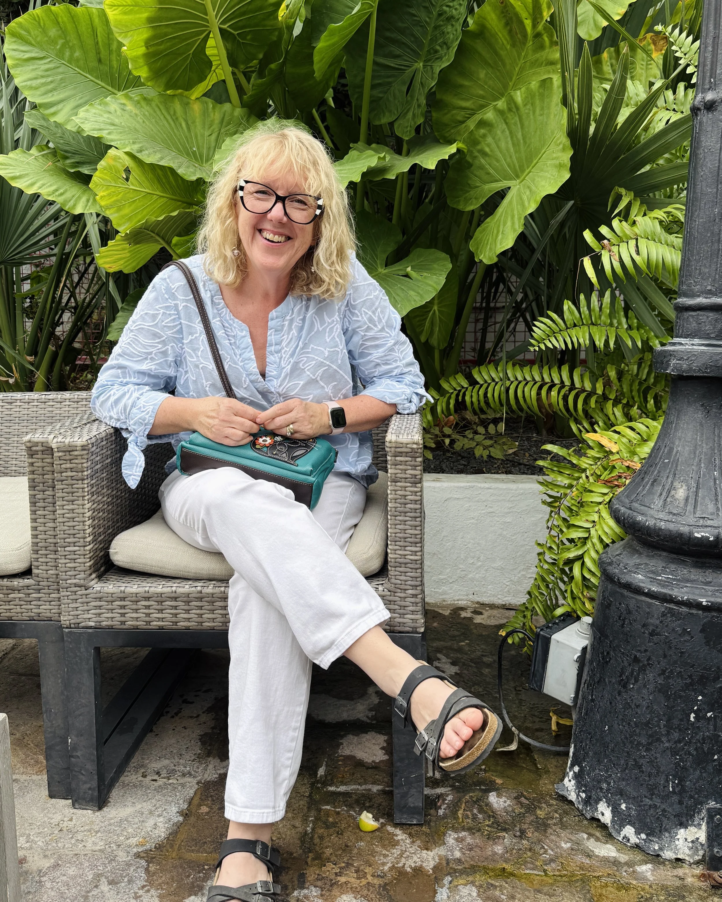 A smiling woman with blonde hair, glasses, and a smartwatch sitting on a wicker bench outdoors among large green leafy plants, wearing a light blue top, white pants, and black sandals.