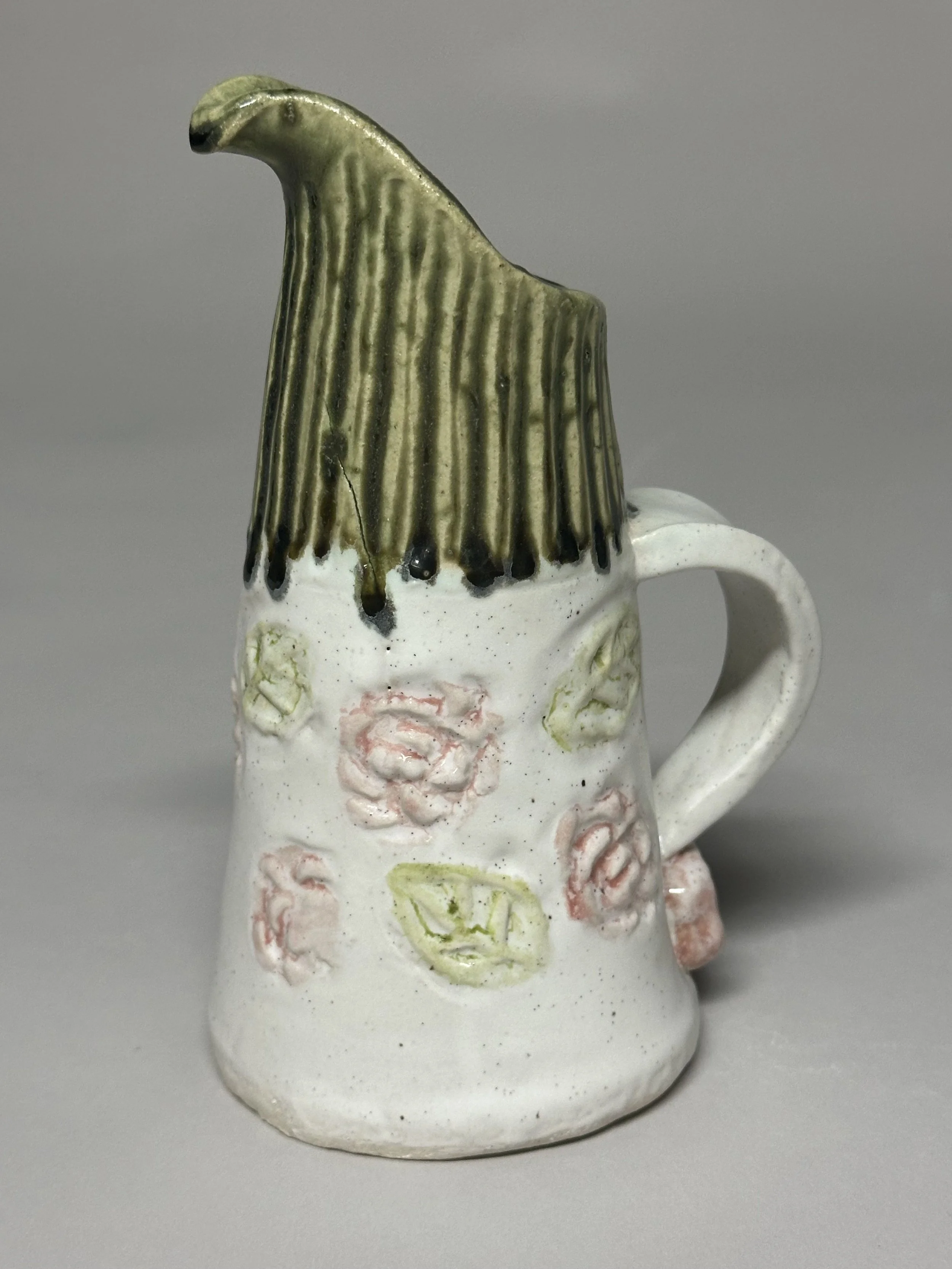 A small, cute pitcher glazed with Santorini Green, soft oatmeal, and touches of pink and green, finished with handmade stamps and a hand‑carved top.