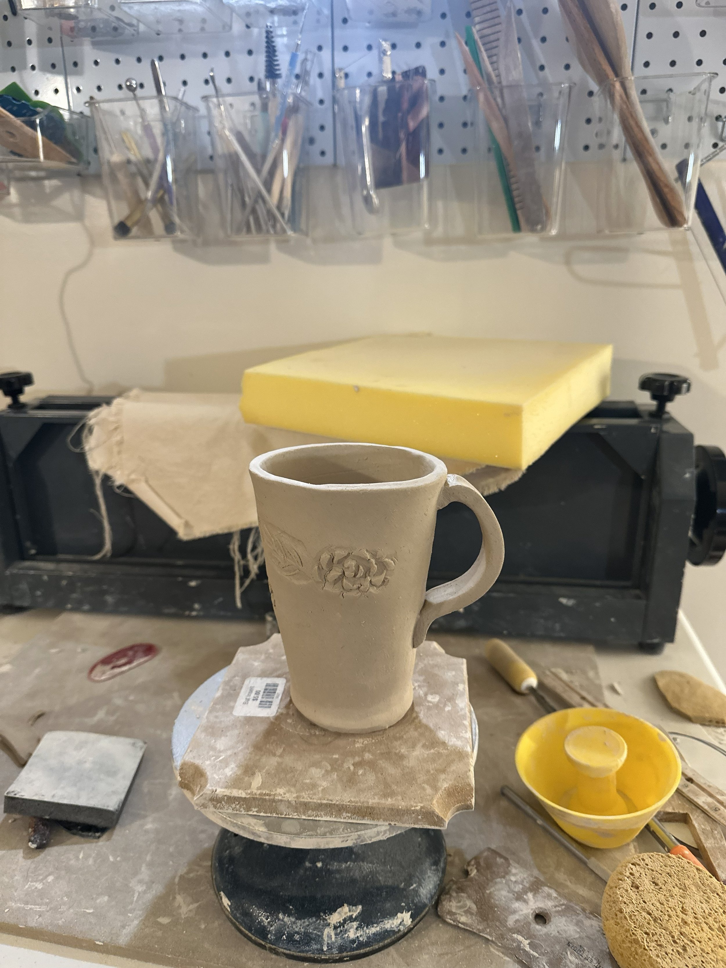 Ceramic mug with floral design in progress on a sculpting wheel, surrounded by pottery tools, sponge, and work materials in a pottery studio.