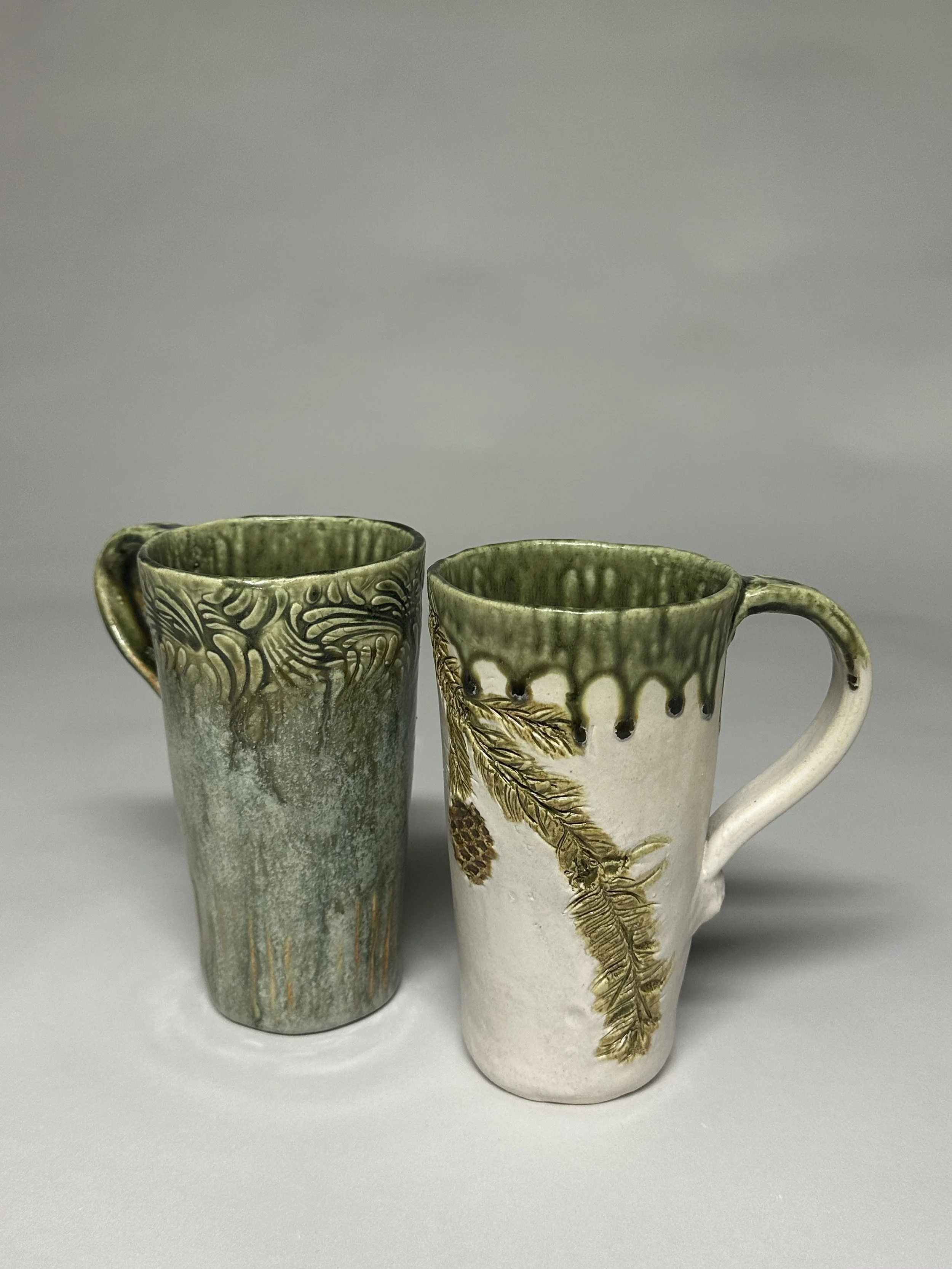Green mug glazed with a combination of eight layered glazes, creating depth, movement, and a richly textured finish. Pinecone hand‑built mug glazed with soft oatmeal and Santorini green glazes, finished with handmade stamps.