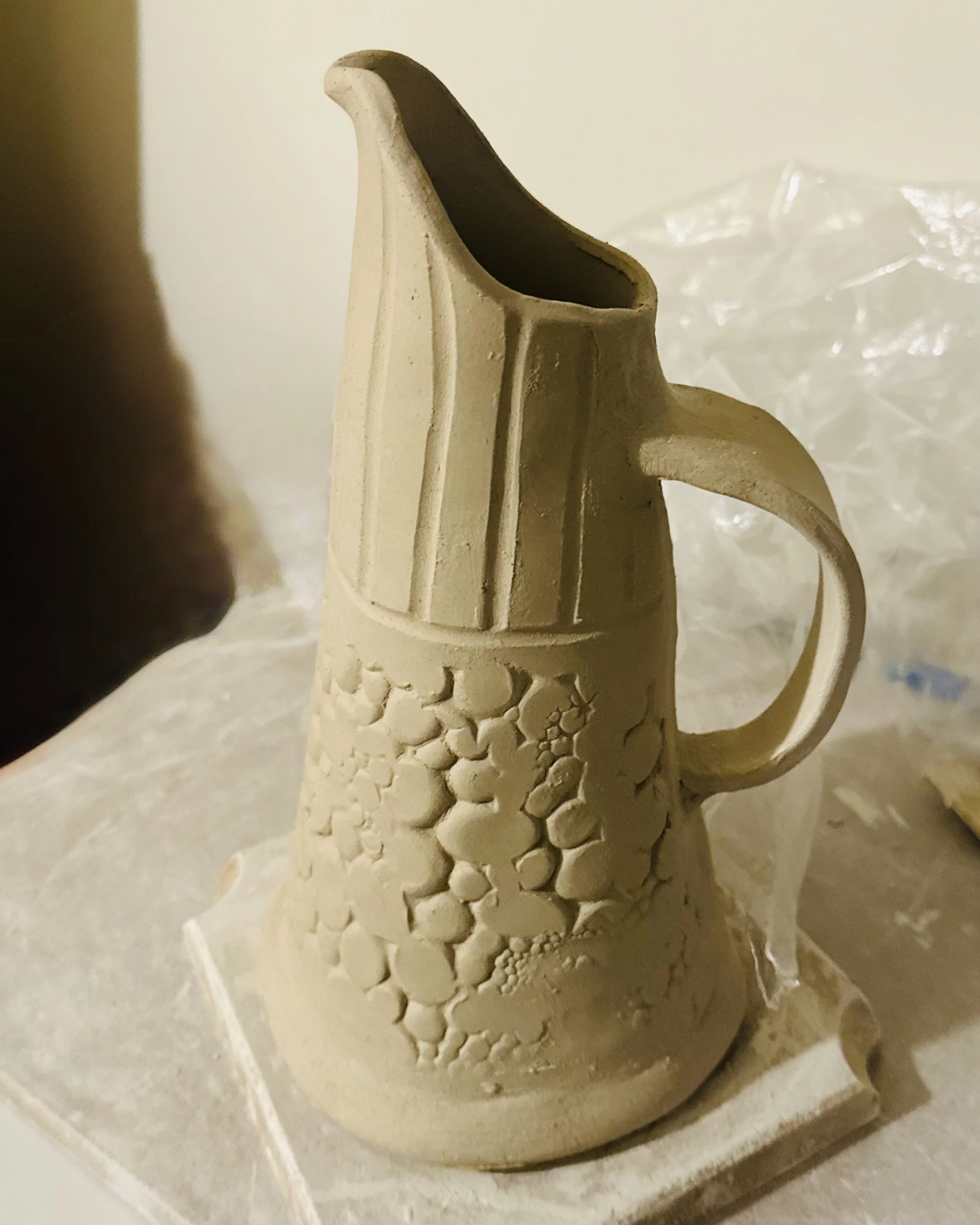 Unfinished ceramic pitcher with textured surface and oval handle, resting on a padded surface.