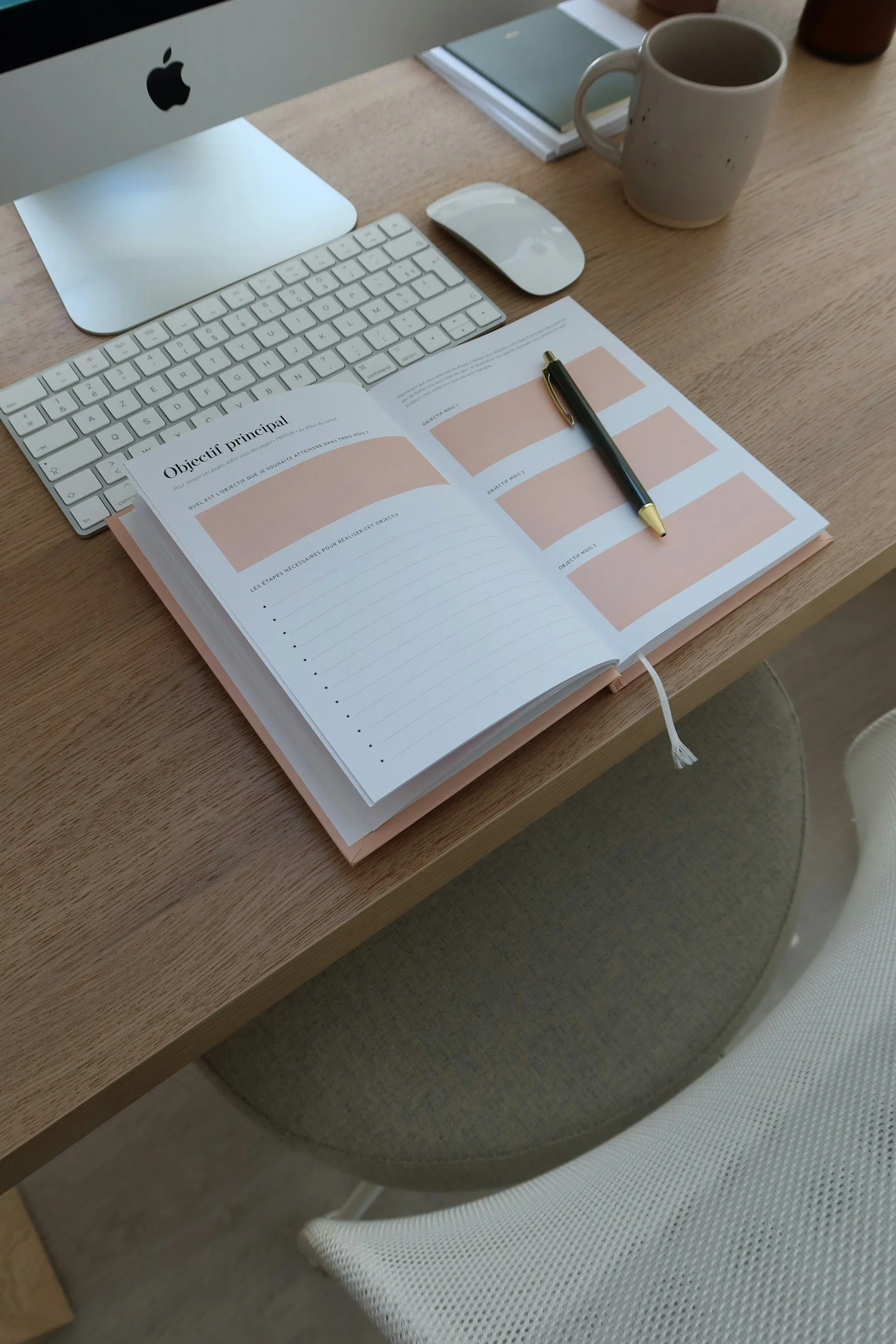 A desk with an iMac computer, wireless keyboard and mouse, an open planner with a pen, a coffee mug, and a notebook.