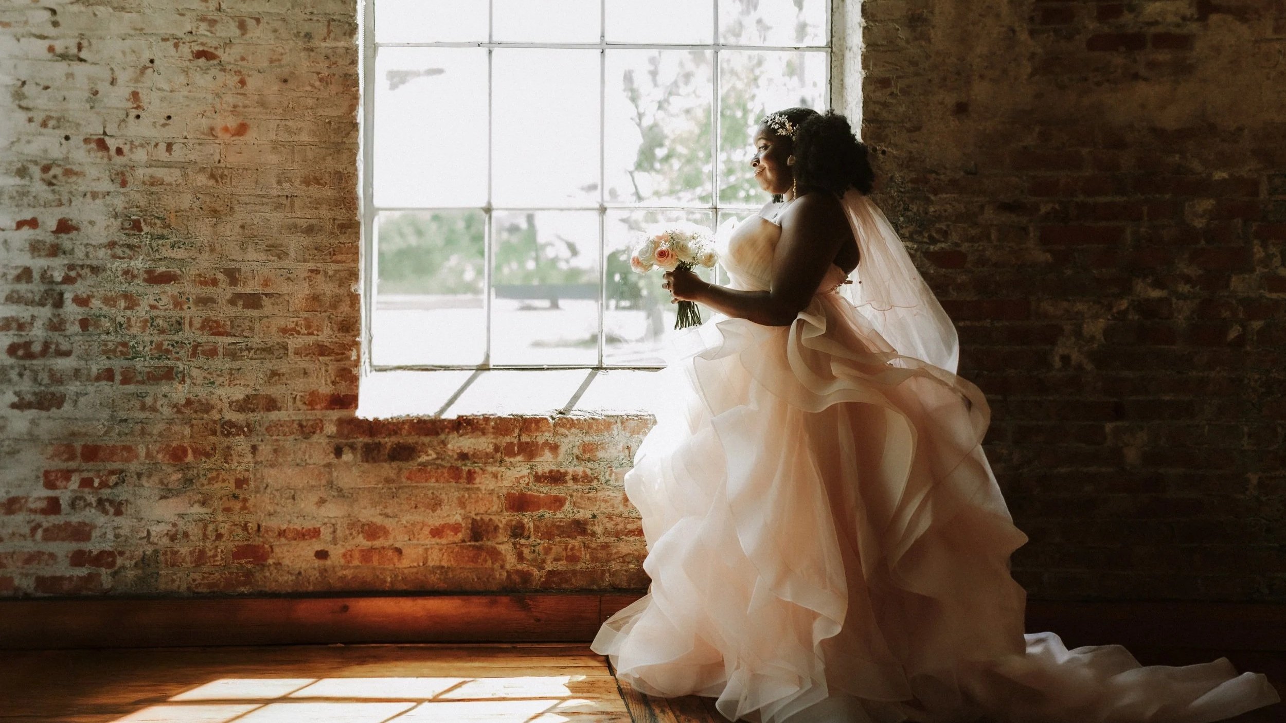 A bride wearing a light-colored wedding gown and veil holding a bouquet of flowers, standing near a large industrial-style window with brick walls and wooden floor, during daytime. Wedding Photos taken at The Venue in Hattiesburg, MS. Editorial