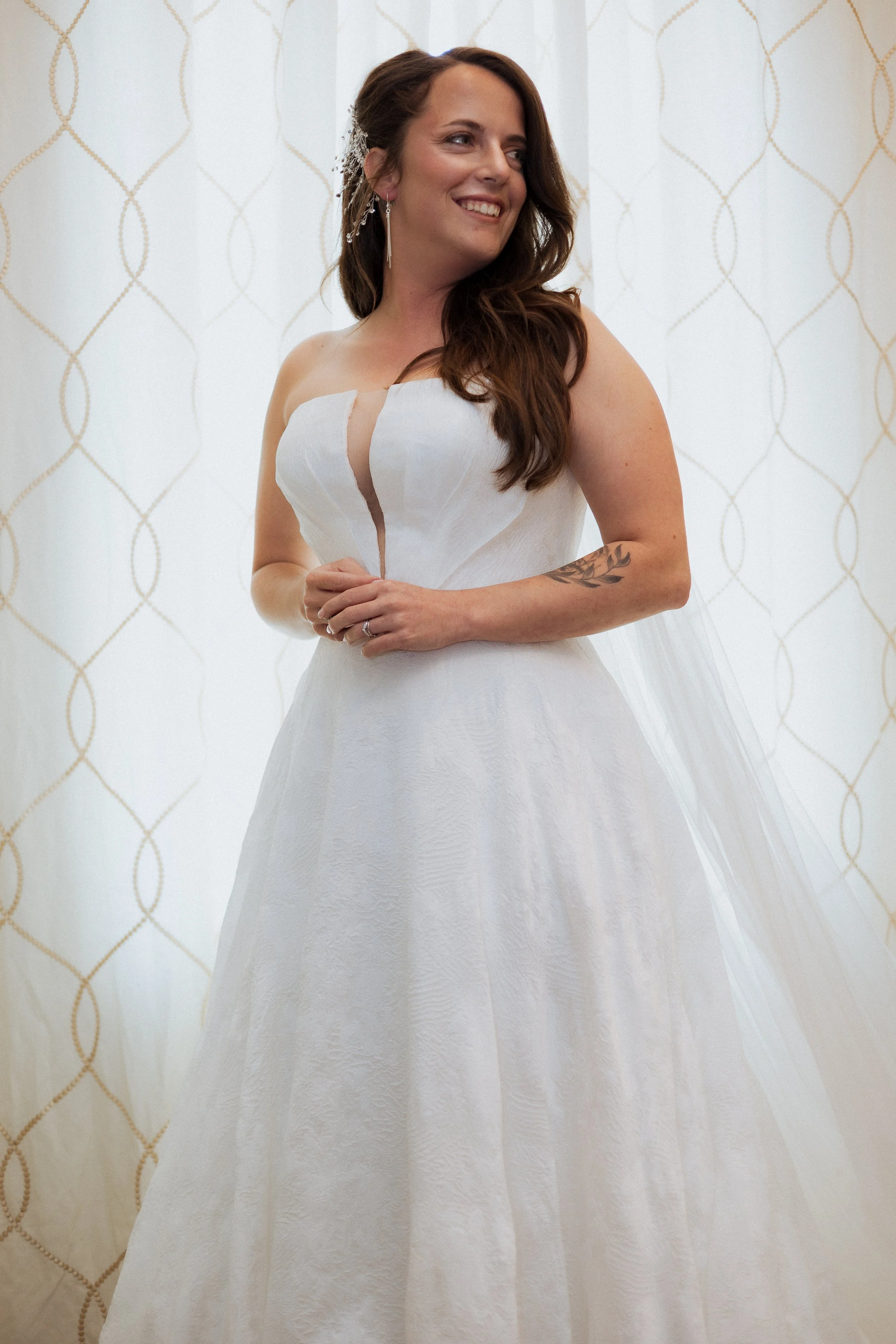 A bride in a white wedding dress with a slit, standing indoors in front of a patterned curtain, smiling and looking to the side. Bainsfield Farm Wedding Venue