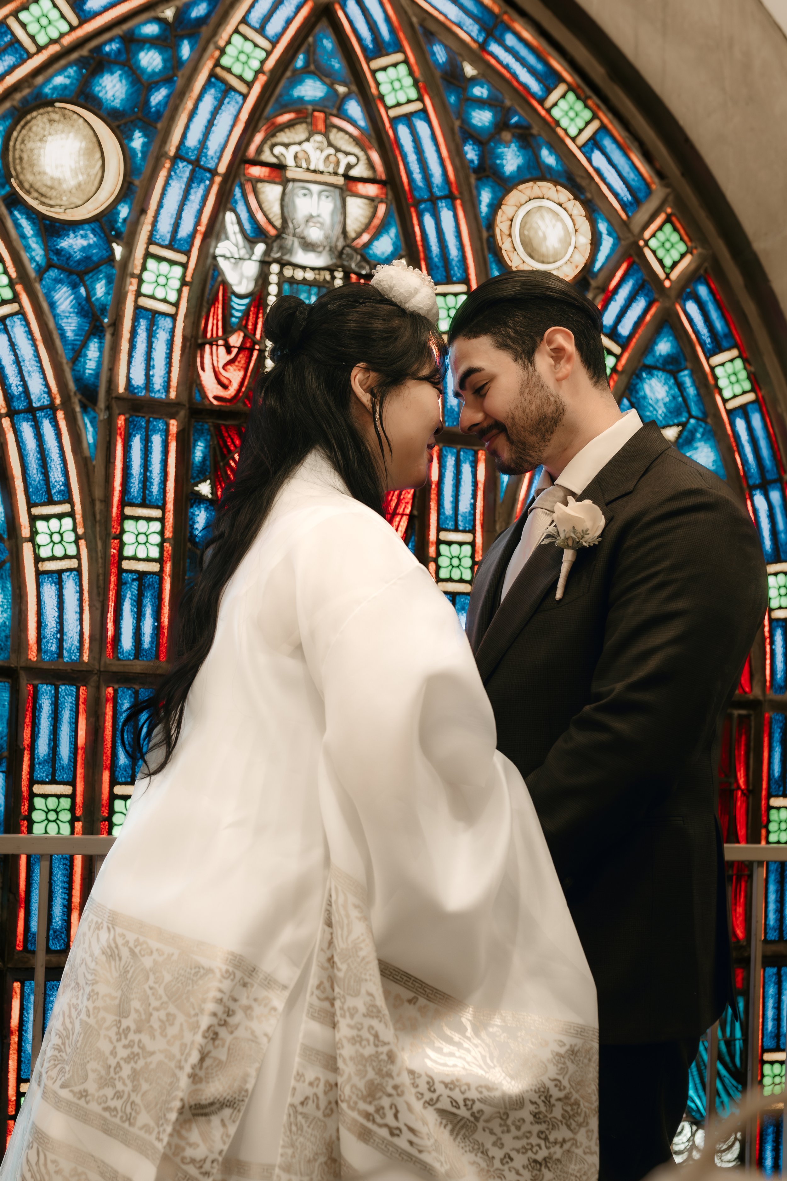 A bride and groom stand close together in front of a stained glass church window. The bride has long dark hair styled in loose waves, wearing a white wedding dress with intricate lace details at the hem. The groom has dark hair, a beard, and is dress