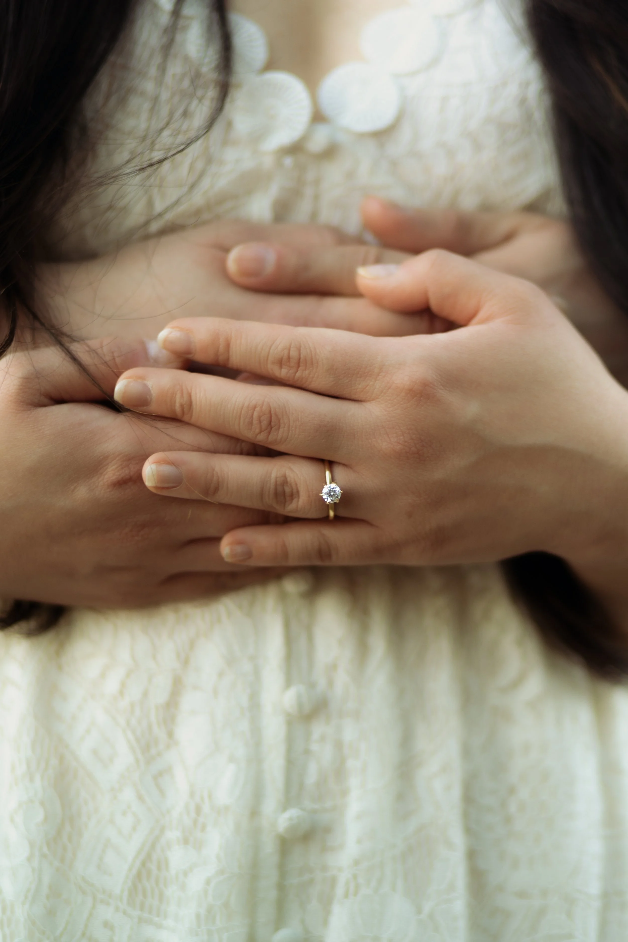 Close-up of woman wearing a yellow dress with a diamond engagement ring on her finger, with hands placed over her chest.