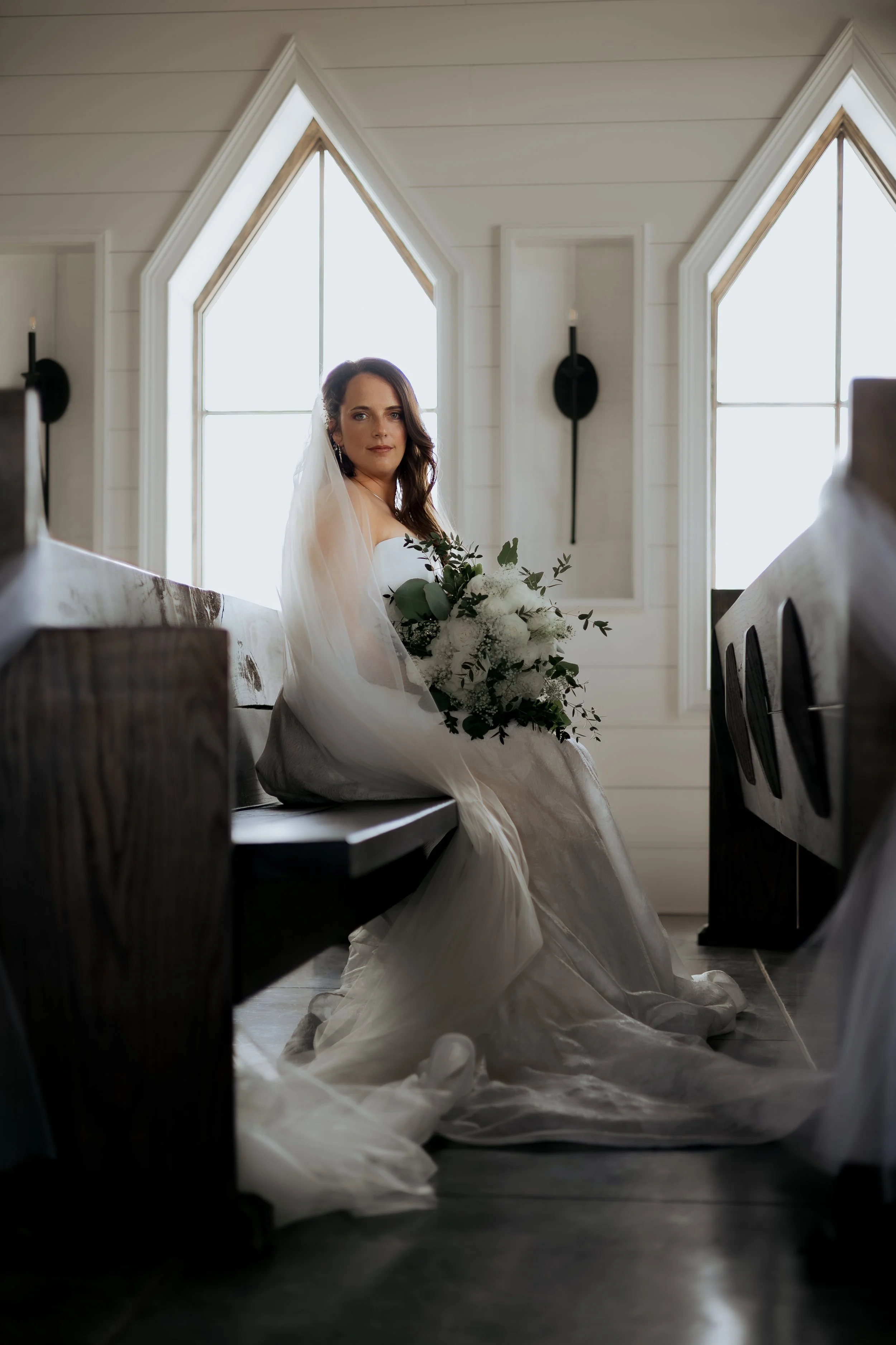 Bride sitting in a church, holding a bouquet of white flowers, with large windows and candle sconces in the background. Bainsfield Farm in Ethelsville, AL.
