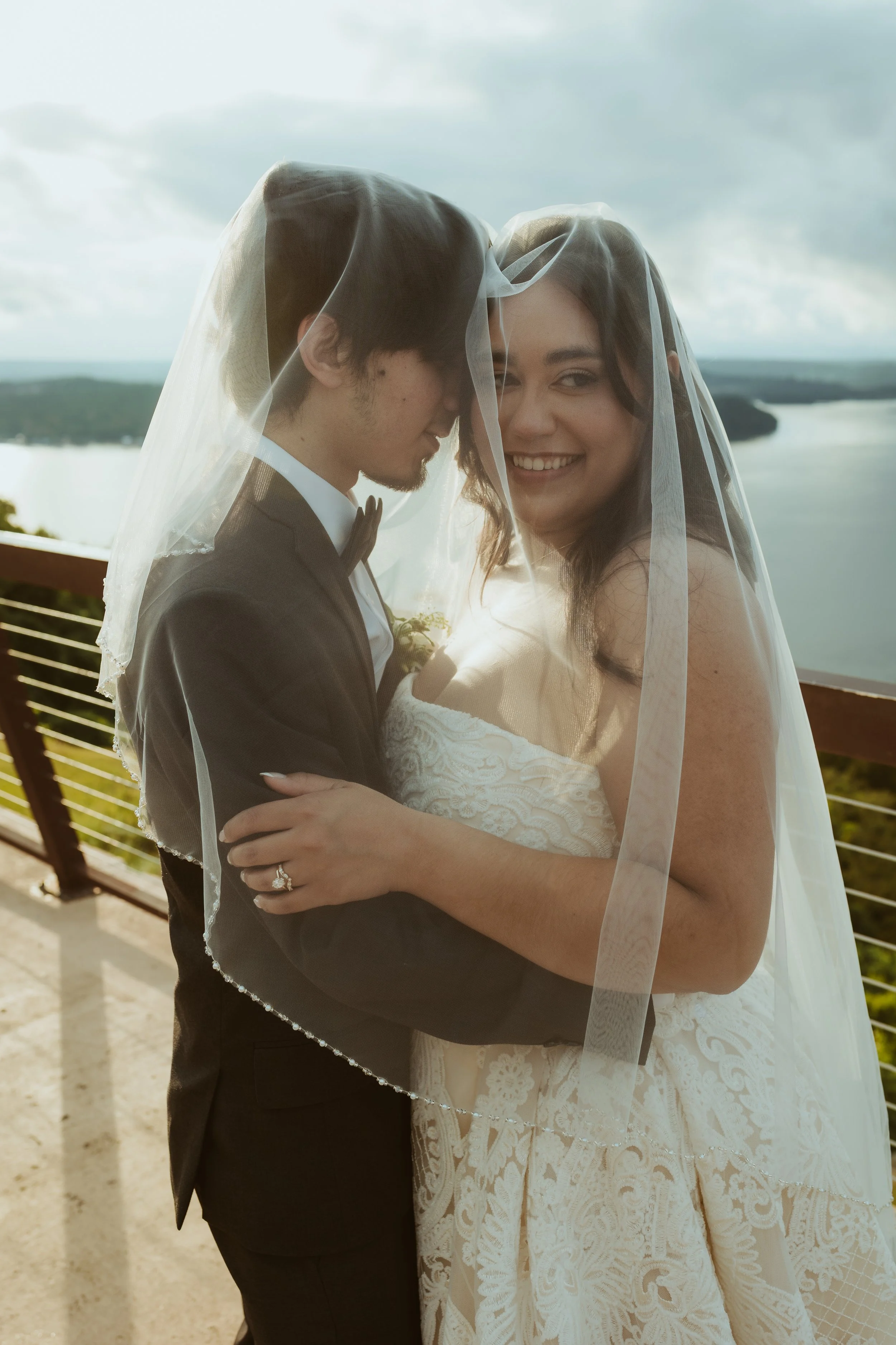 A newlywed couple standing on an outdoor balcony, with a view of a body of water and landscape in the background, both wearing wedding attire and veils, embrace and smile. Guntersville, AL Weddings. Wedding photographer in guntersville, al.