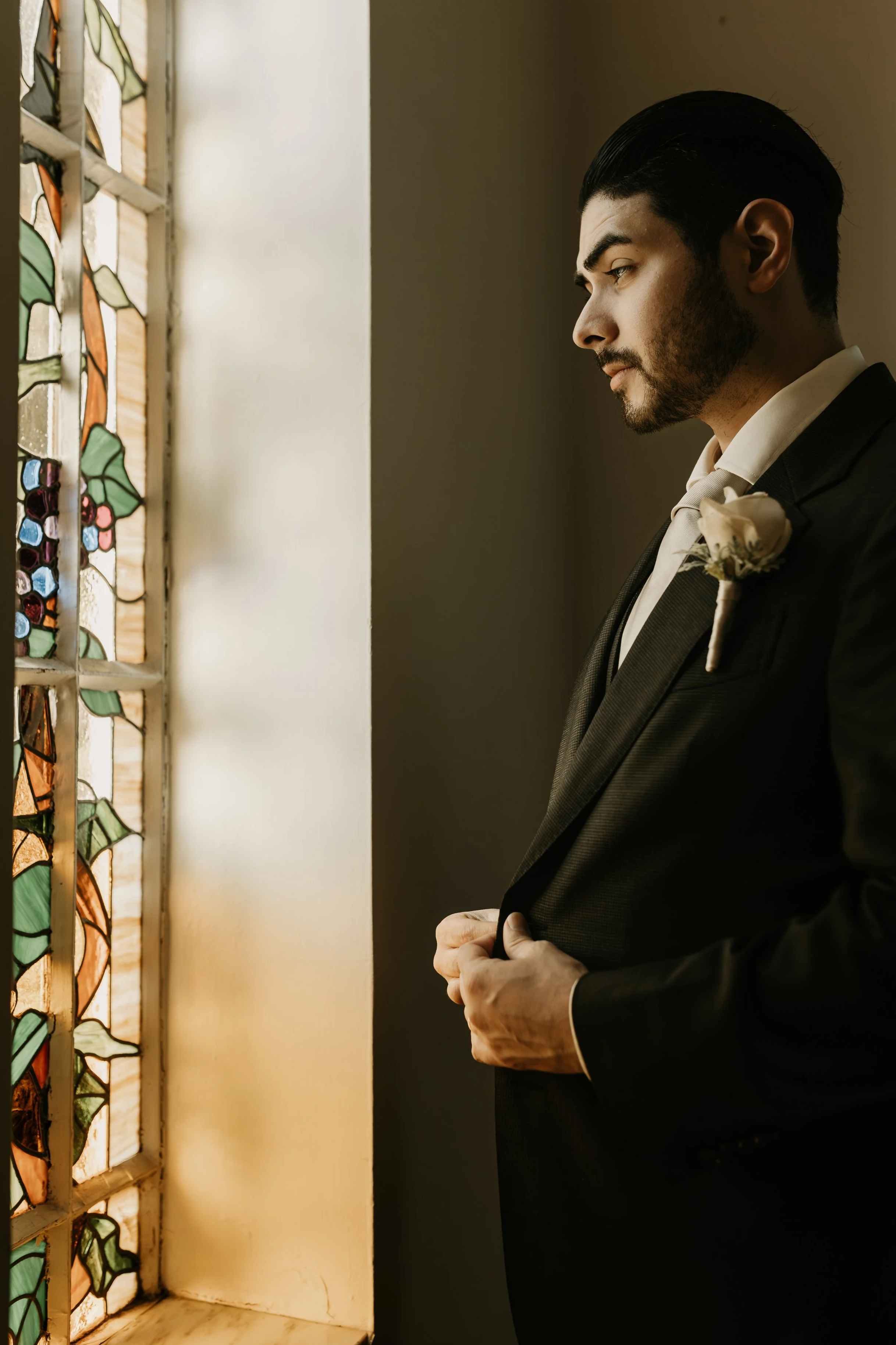A groom in a black suit looking out a stained glass window, standing indoors with a white boutonnière on his lapel.