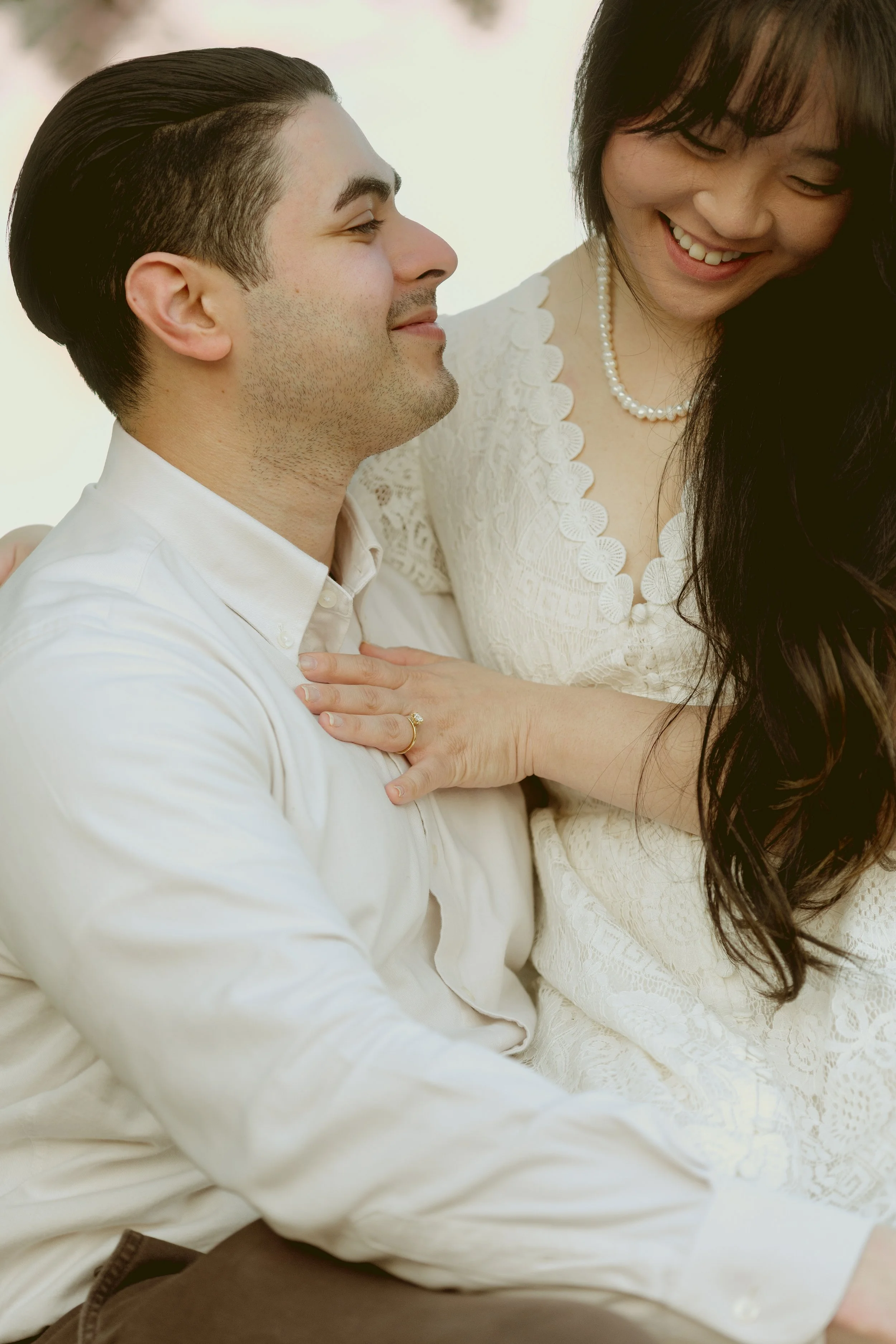 A man and woman smiling and looking at each other, with the woman touching the man's chest, indoors with a bright background.