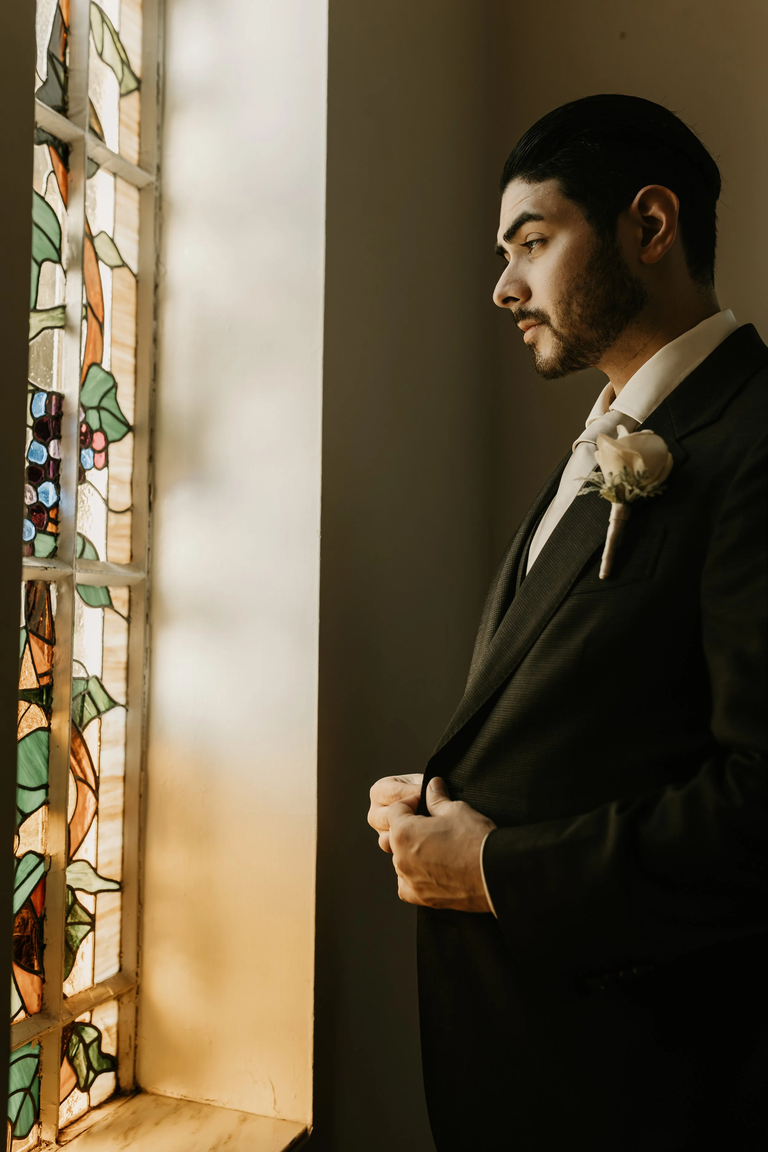 Man in a tuxedo with a boutonniere looking out a stained glass window. Alabama Wedding Photographer with a editorial style.