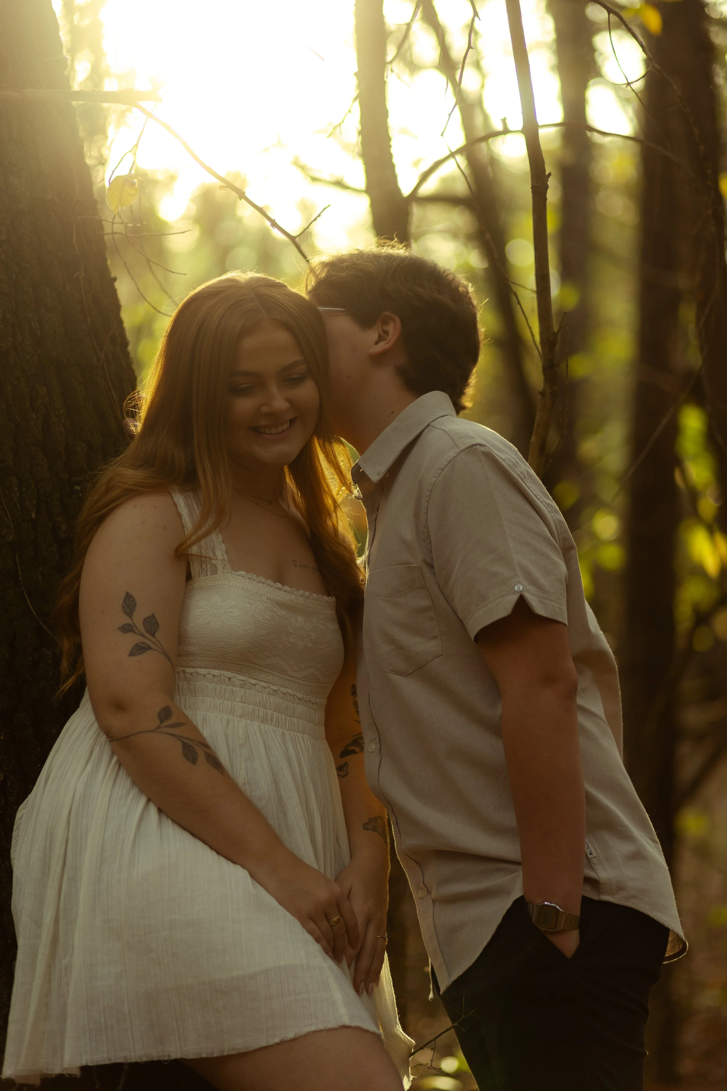 A couple shares a tender moment in a sunlit forest, with the woman smiling and the man whispering in her ear.