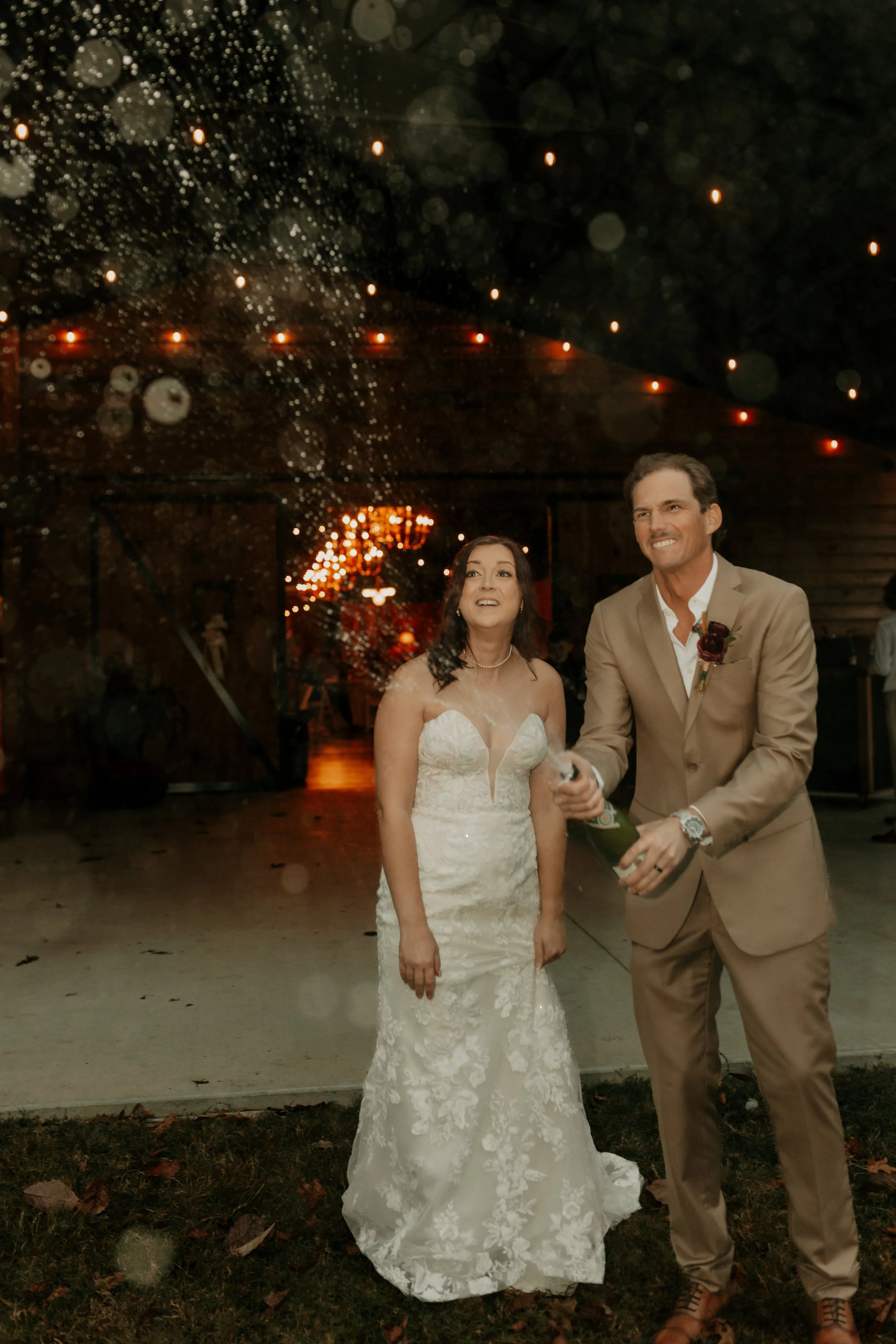 A bride and groom celebrating under a confetti shower at their wedding reception. The bride is wearing a white lace strapless wedding gown, and the groom is in a tan suit with a boutonniere. They are smiling and holding a champagne bottle.
