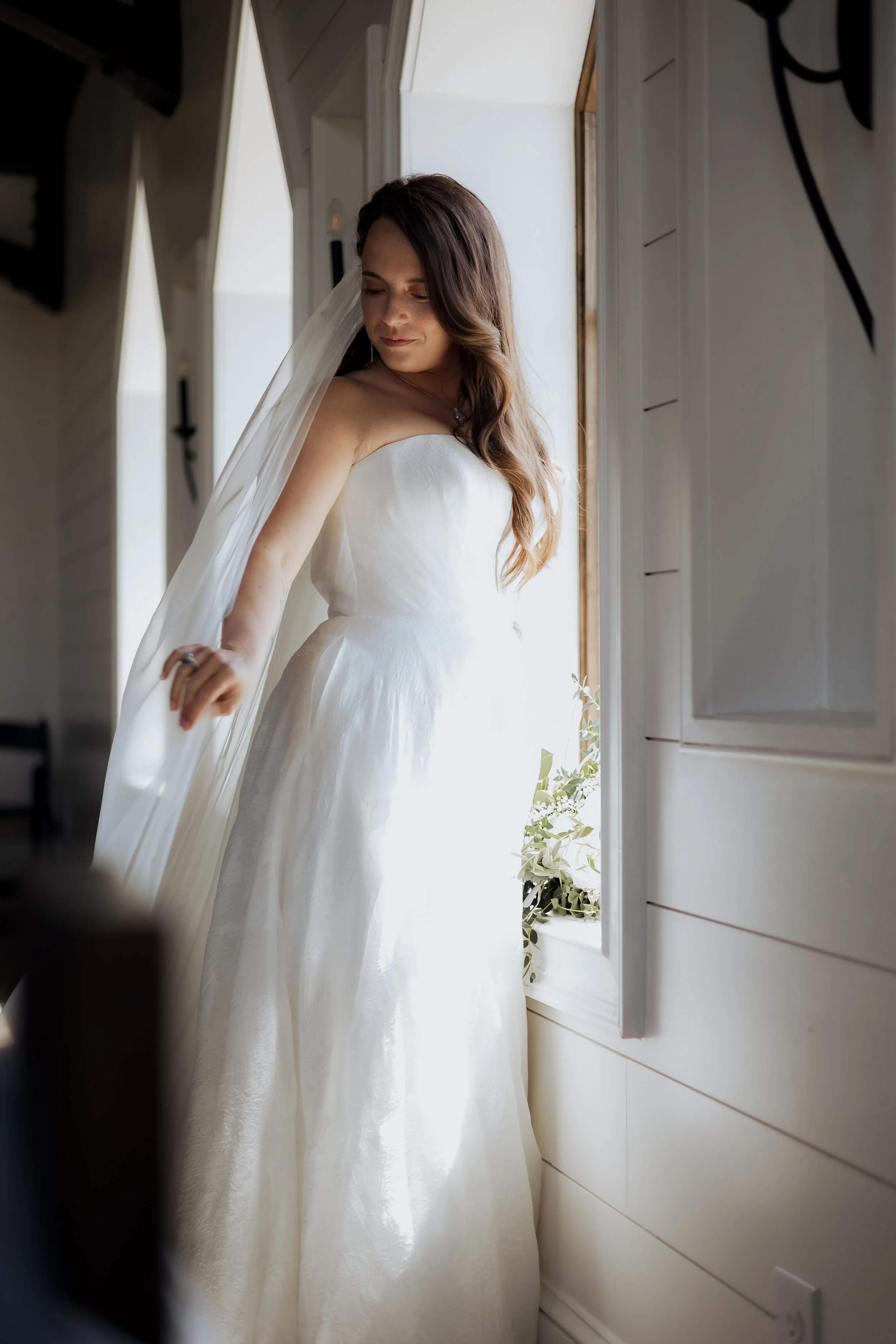 A bride in a strapless white wedding dress standing by an open window, with sunlight illuminating her profile as she looks downward.