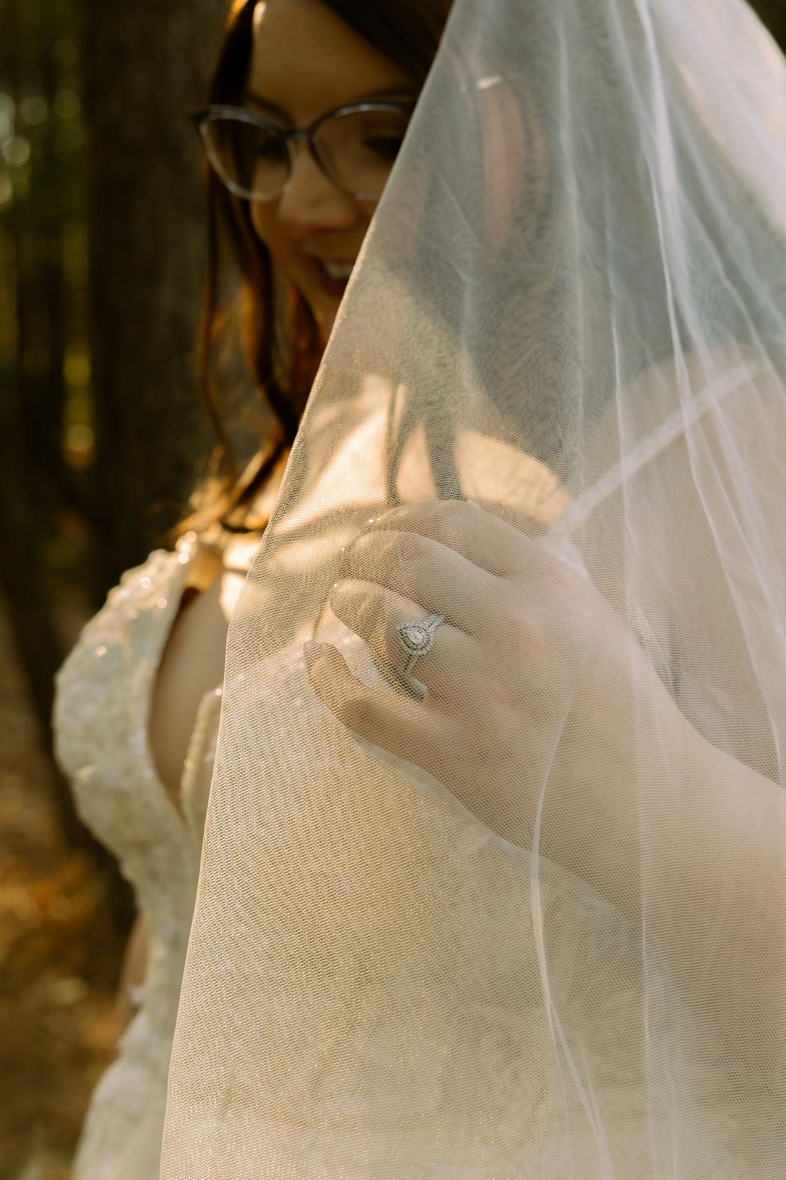 A woman with glasses and red hair, wearing a wedding dress, is holding a veil and smiling outdoors.