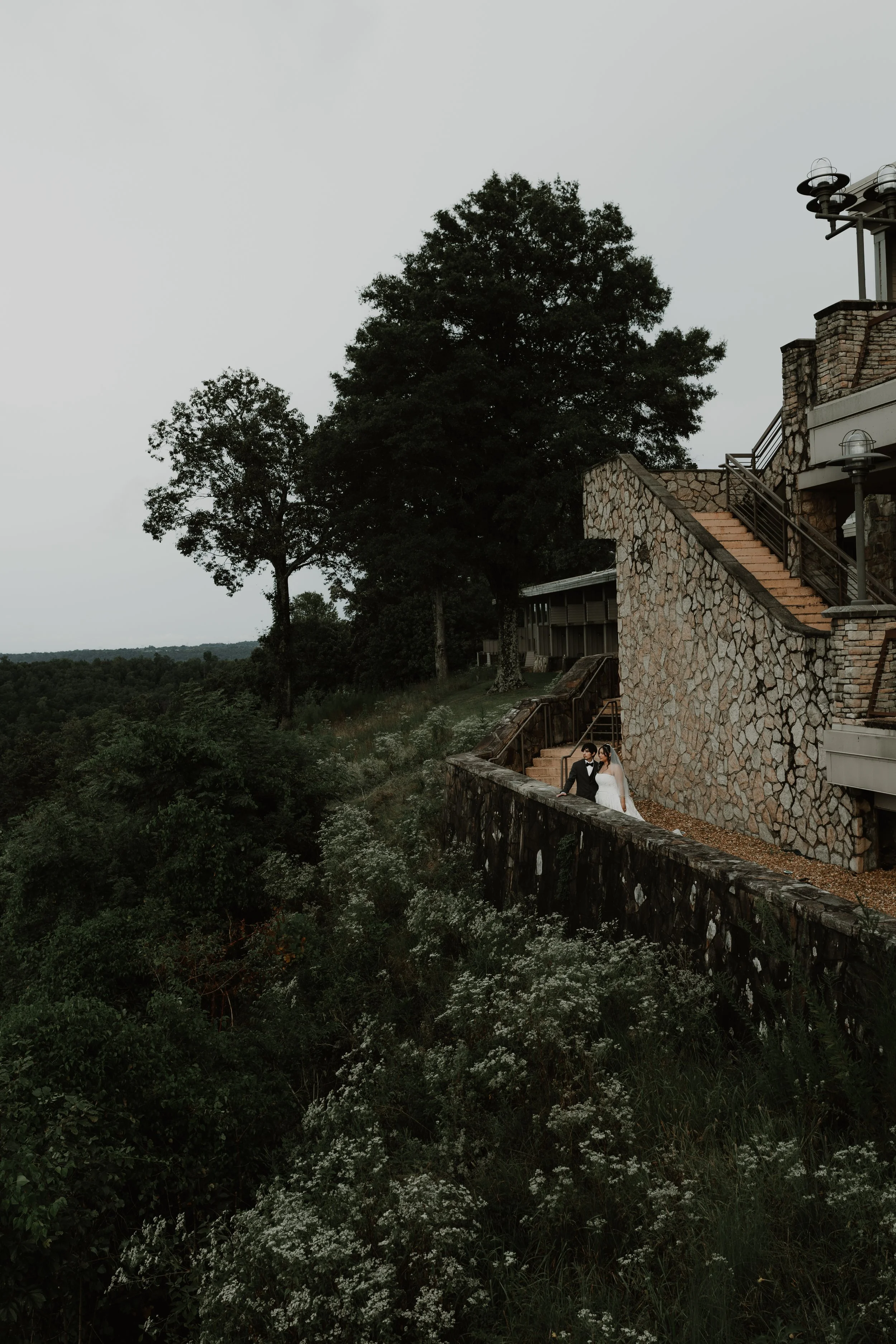 A bride and groom walking down a stone walkway on a hillside with greenery and trees landscape, outside a stone building under overcast sky. Editorial Wedding Photography in Alabama. 