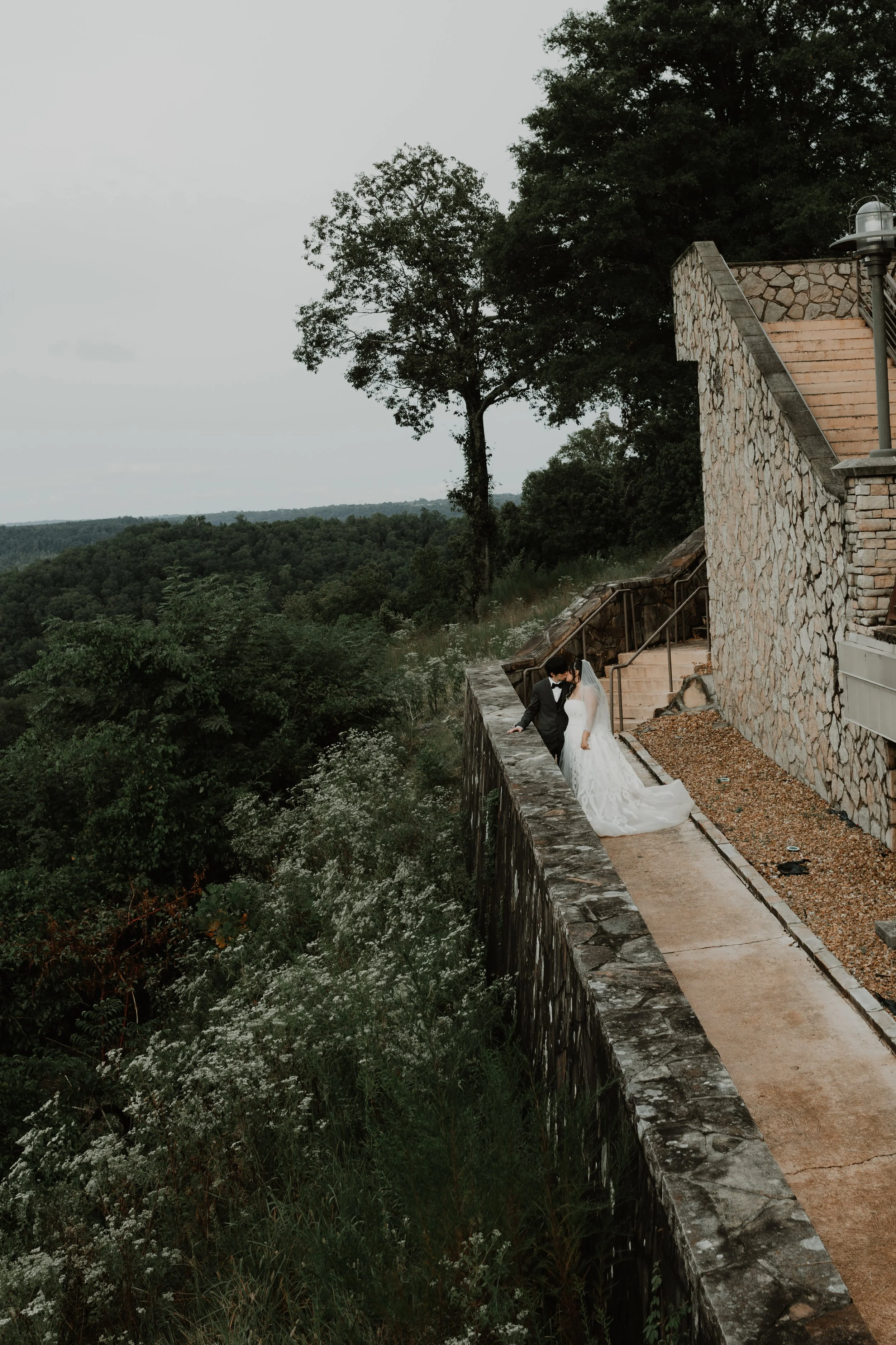 A bride and groom standing on a stone wall overlooking a forested landscape, with trees and cloudy sky in the background. Guntersville, AL Weddings. Wedding photographer in guntersville, al.
