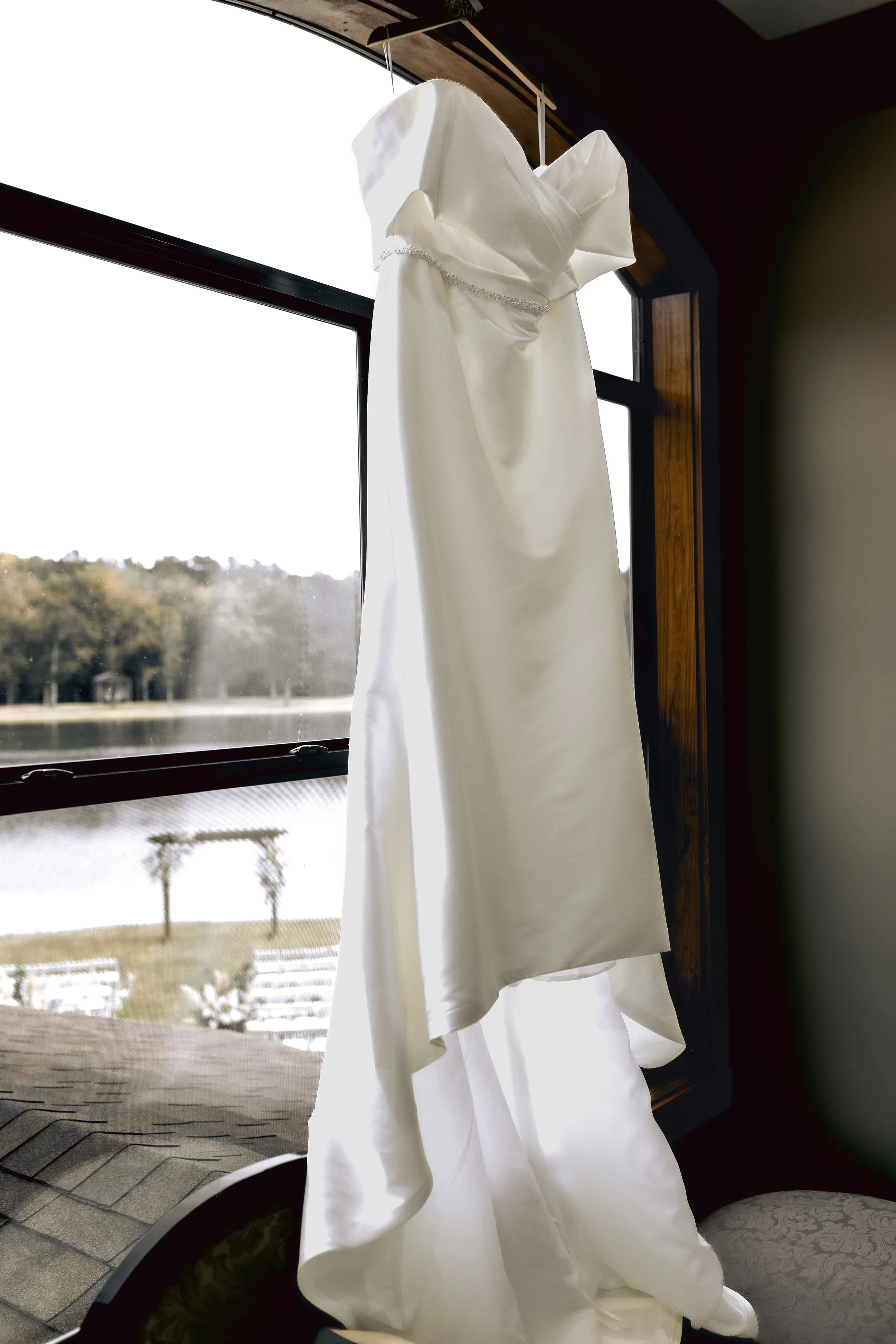 White wedding dress hanging in front of a window with a view of trees and a lake outside. Behind The Pines Wedding Venue. Wedding Photography in Mississippi.