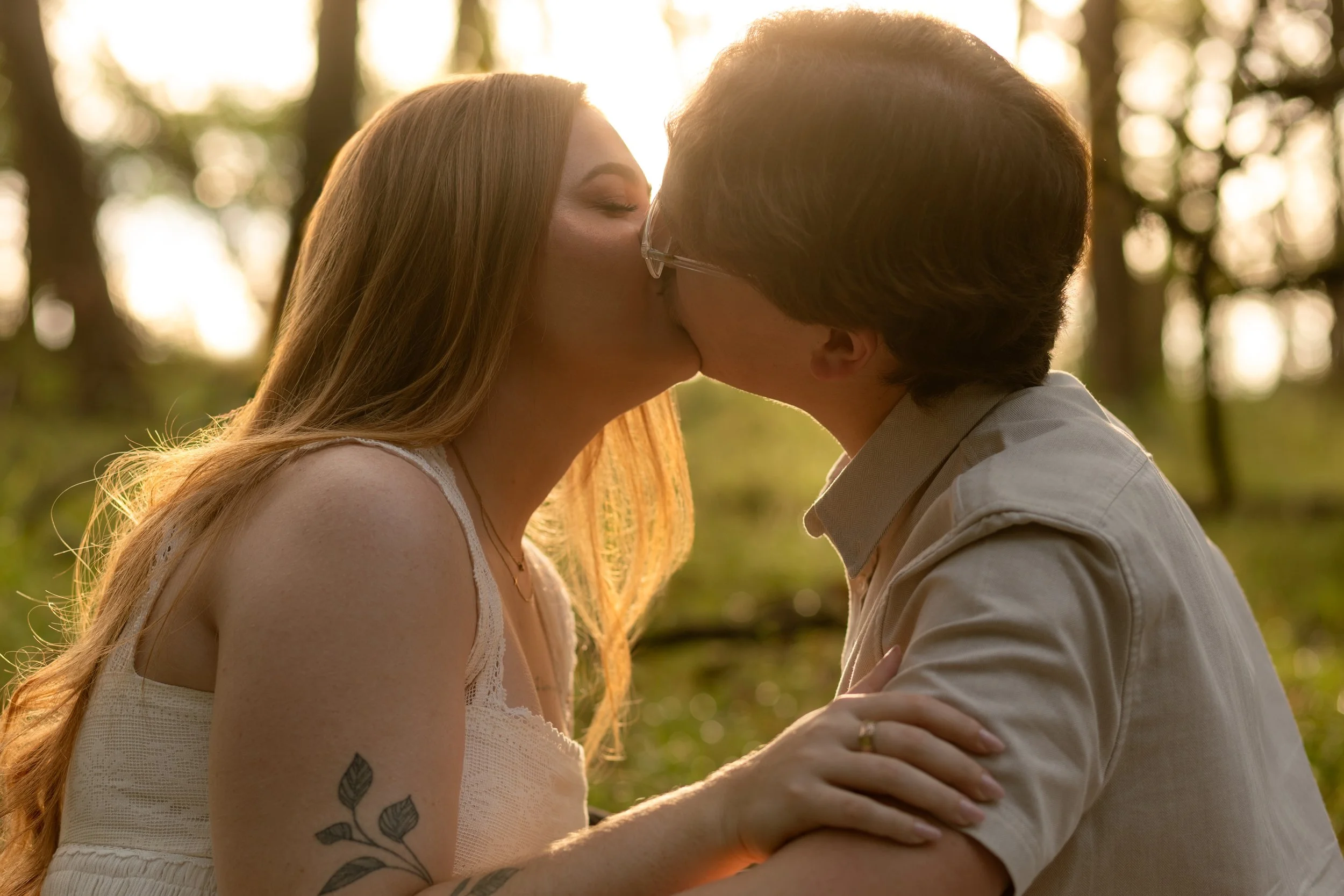 A couple sharing a kiss outdoors during sunset, with trees in the background and sunlight shining through.